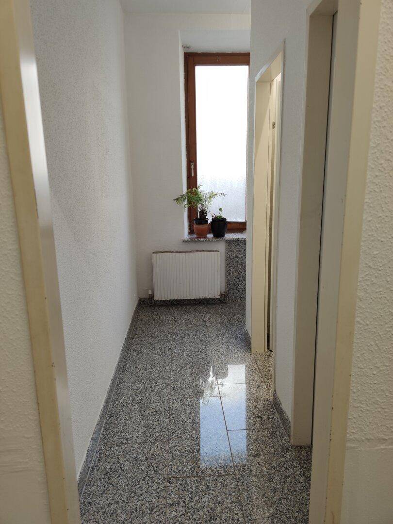 Schmaler Flur mit Steinboden, Heizkörper und einem Fenster am Ende des Ganges.