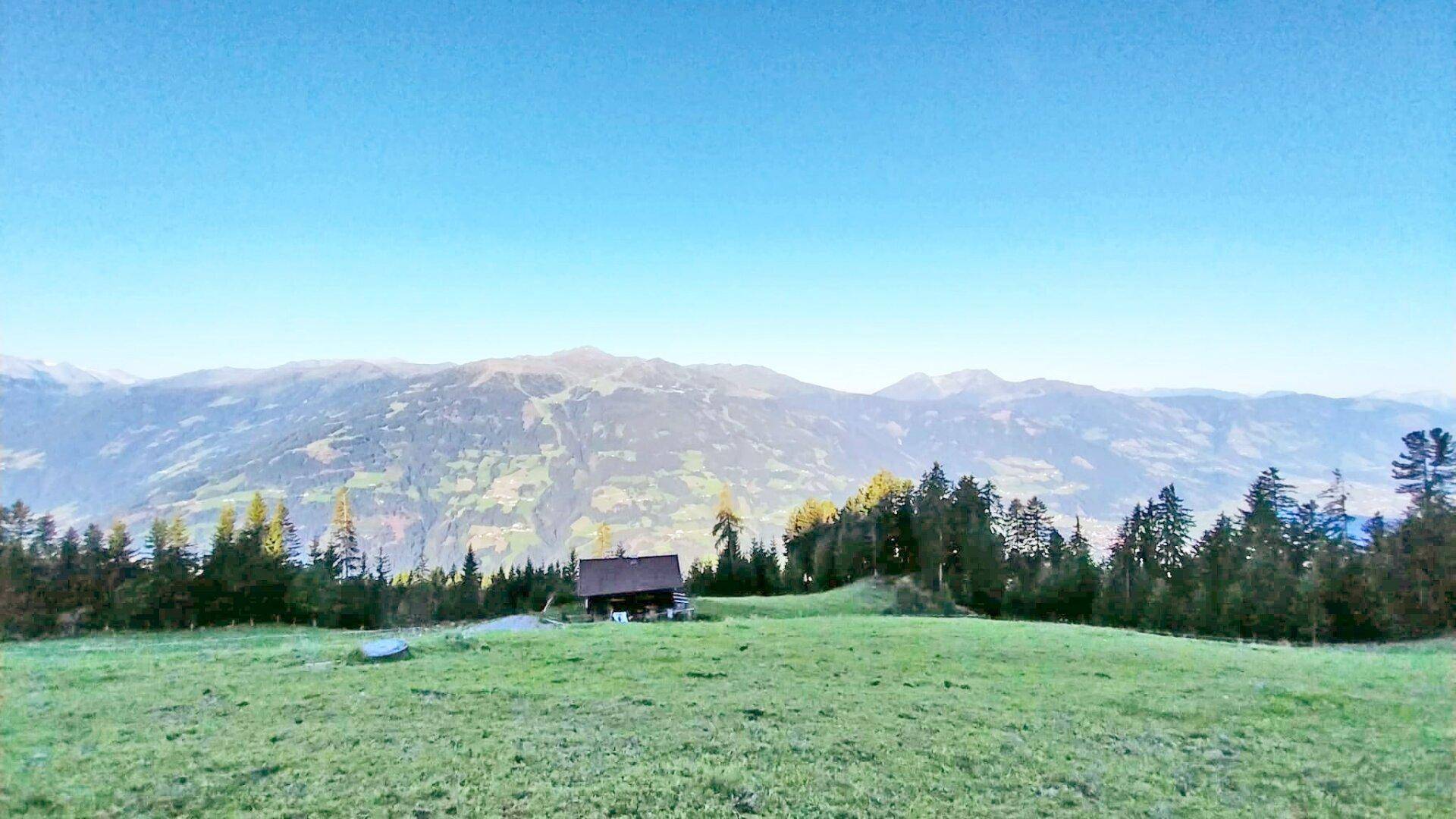 Kleine Holzhütte in der Ferne, eingebettet in eine weite, grüne Berglandschaft unter blauem Himmel.