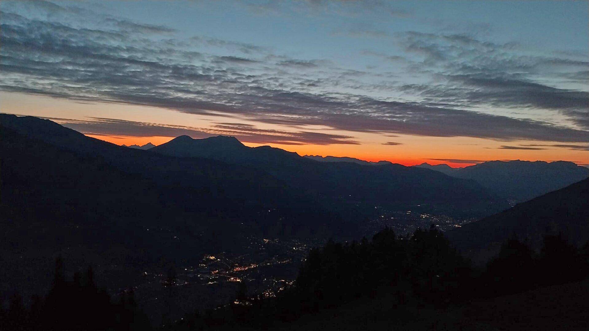 Abenddämmerung über einem beleuchteten Tal mit Bergen im Hintergrund und einem farbigen Horizont.