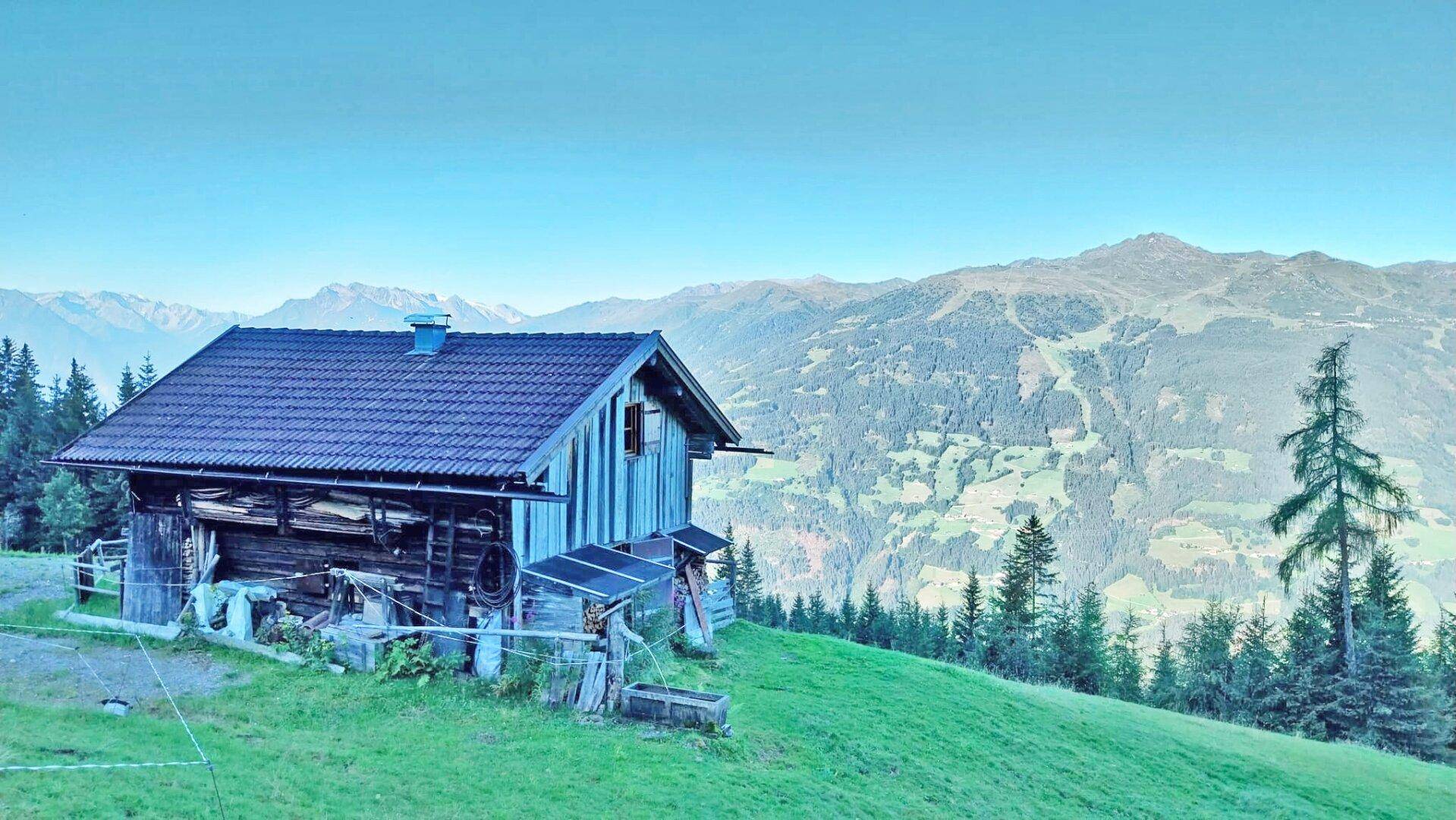 Holzhütte in einer weiten Berglandschaft unter klarem Himmel, umgeben von Wald und Wiesen.