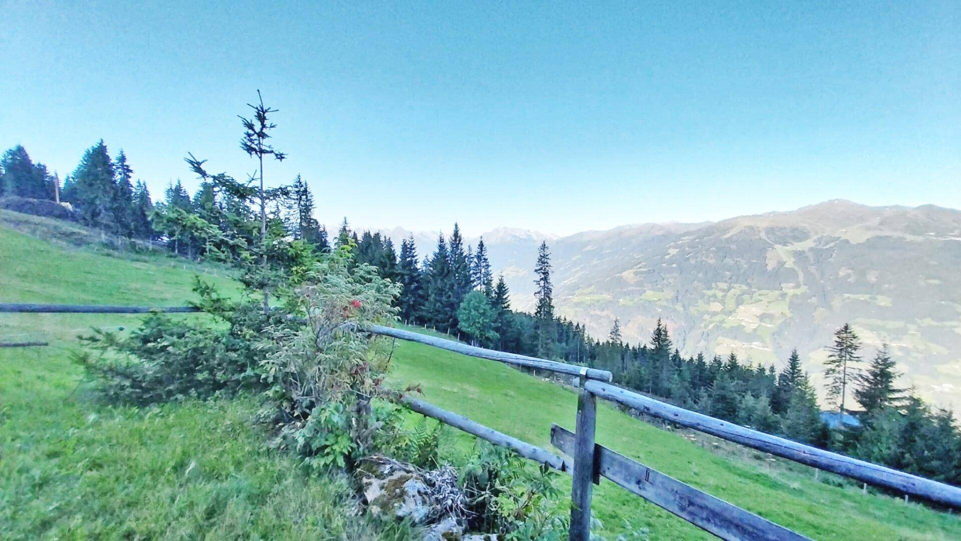 Grüne Wiesen und ein rustikaler Holzzaun mit Blick auf die weite Berglandschaft.