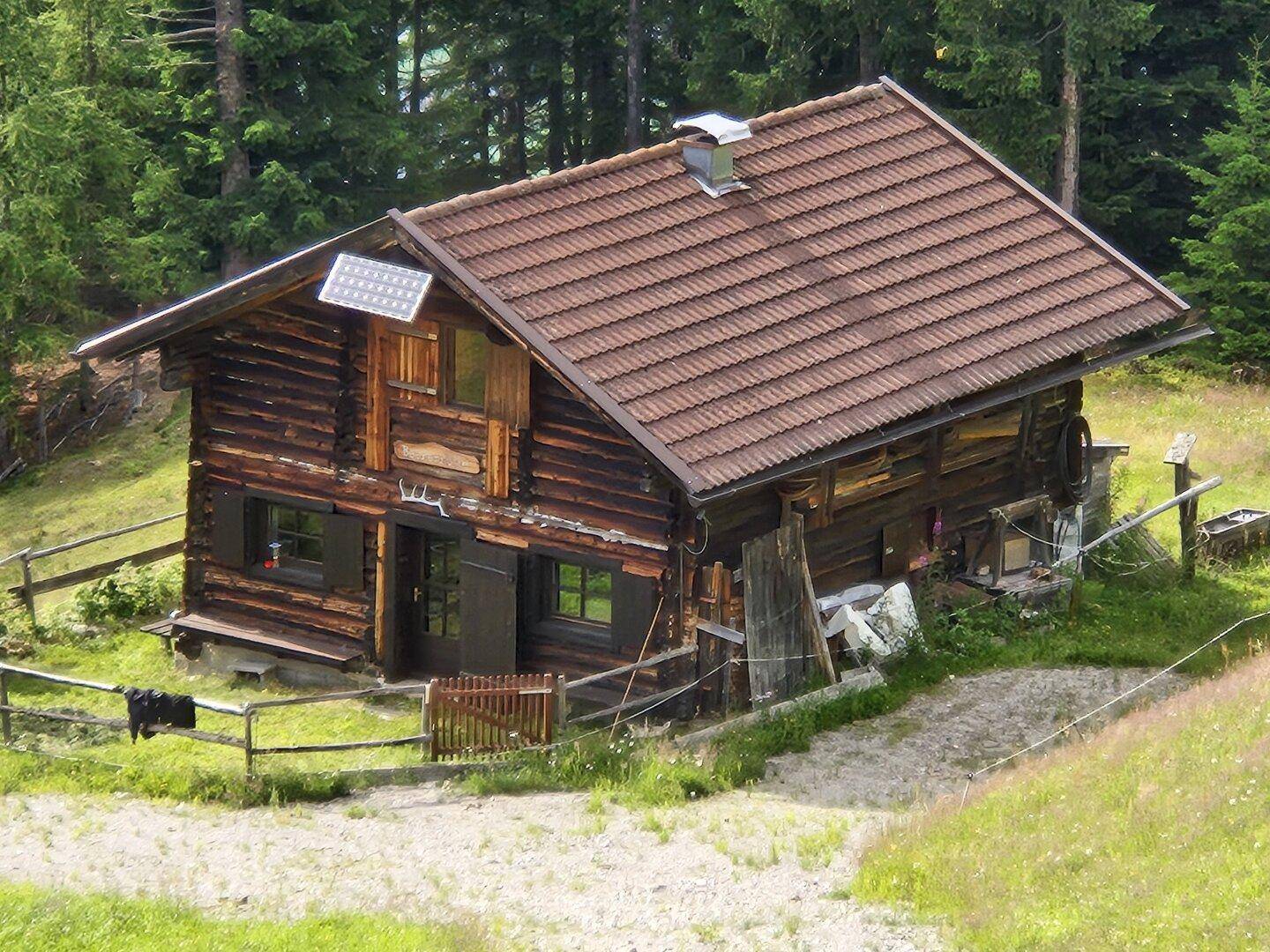 Gemütliche Holzhütte mit Ziegeldach und Solarpaneel, umgeben von grünen Wiesen und Wald.