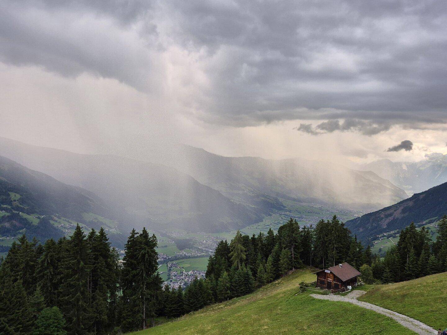 Holzhütte in einem grünen Tal mit Bergen im Hintergrund und Regenwolken am Horizont.