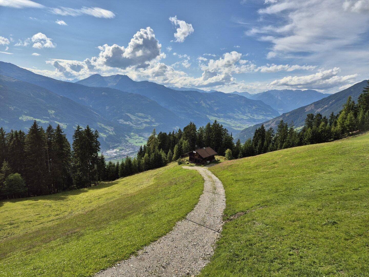 Malerische Holzhütte inmitten einer weiten Berglandschaft mit grünen Wiesen und einem Schotterweg.