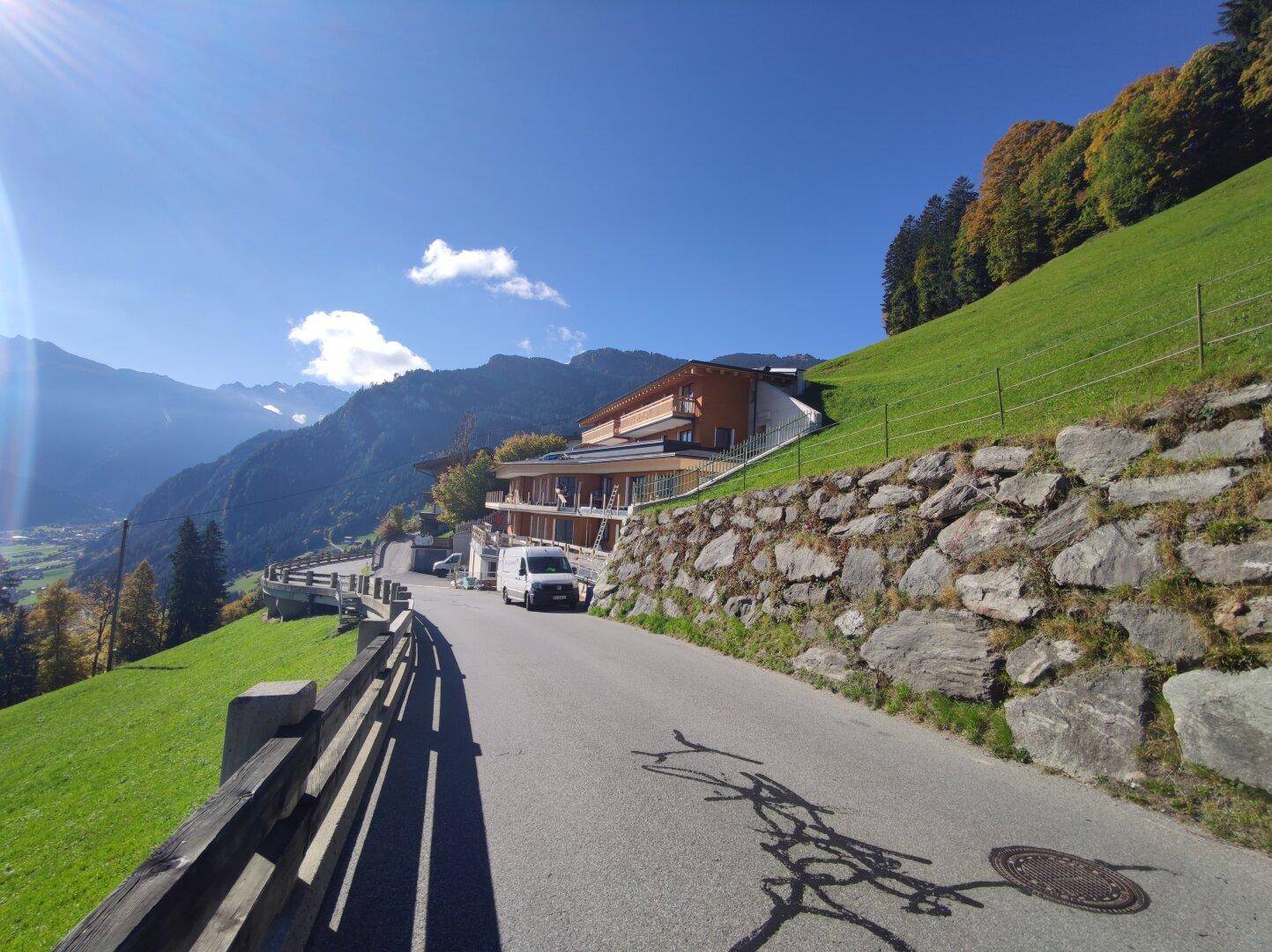 Panoramablick auf das Neubauprojekt in den Bergen mit sonniger Hanglage und Zufahrtsstraße.