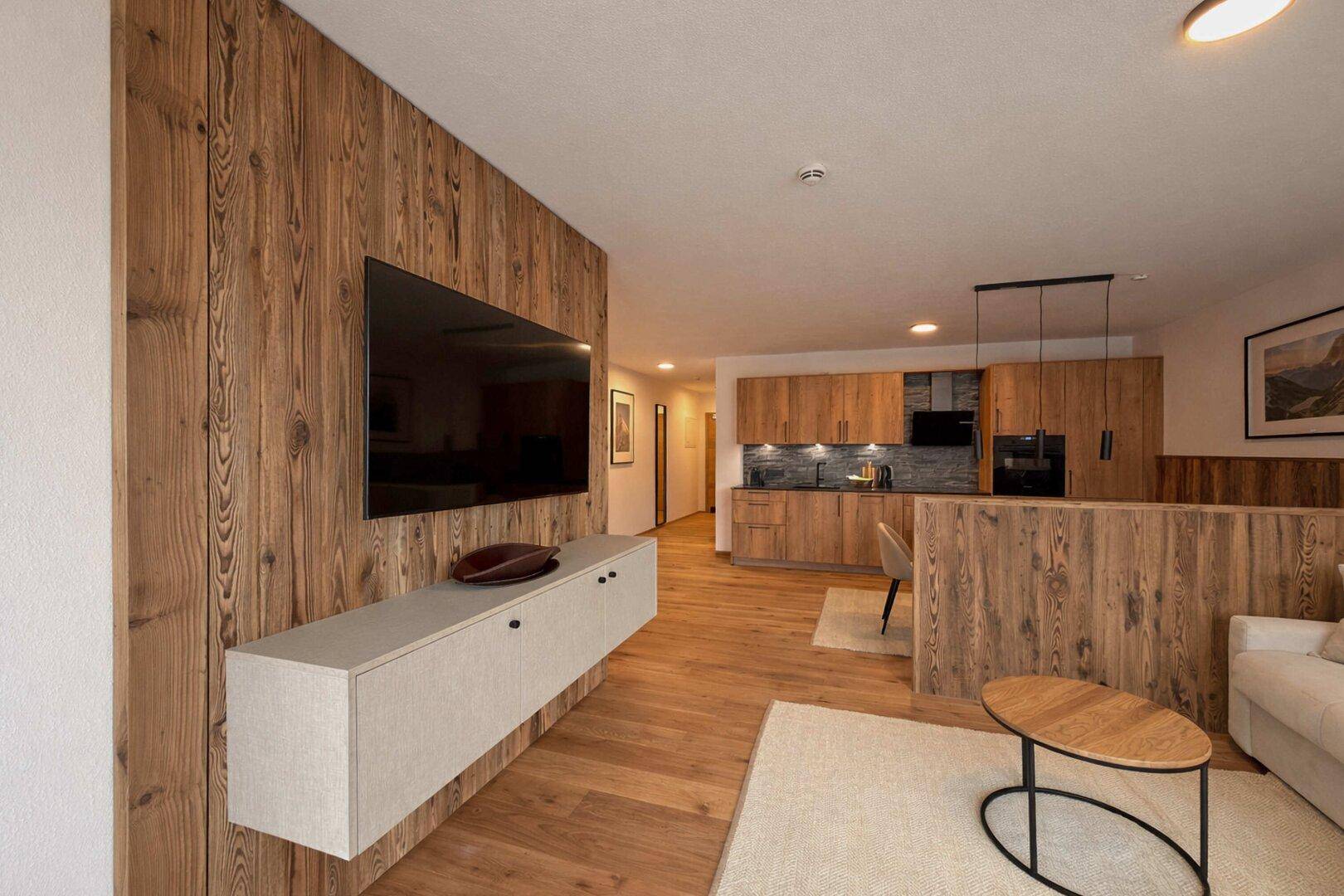 Moderner Wohnbereich mit Holzwand, Flachbildfernseher und offener Küche im Hintergrund.