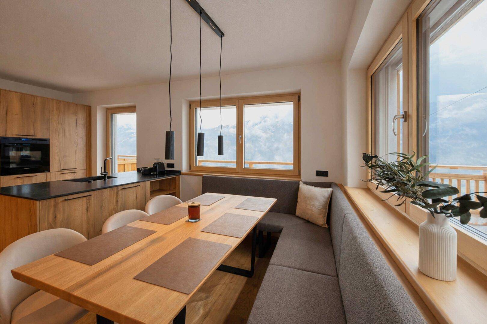 Heller Essbereich mit Holztisch, Sitzbank und Fenstern mit Blick auf die umliegende Berglandschaft.