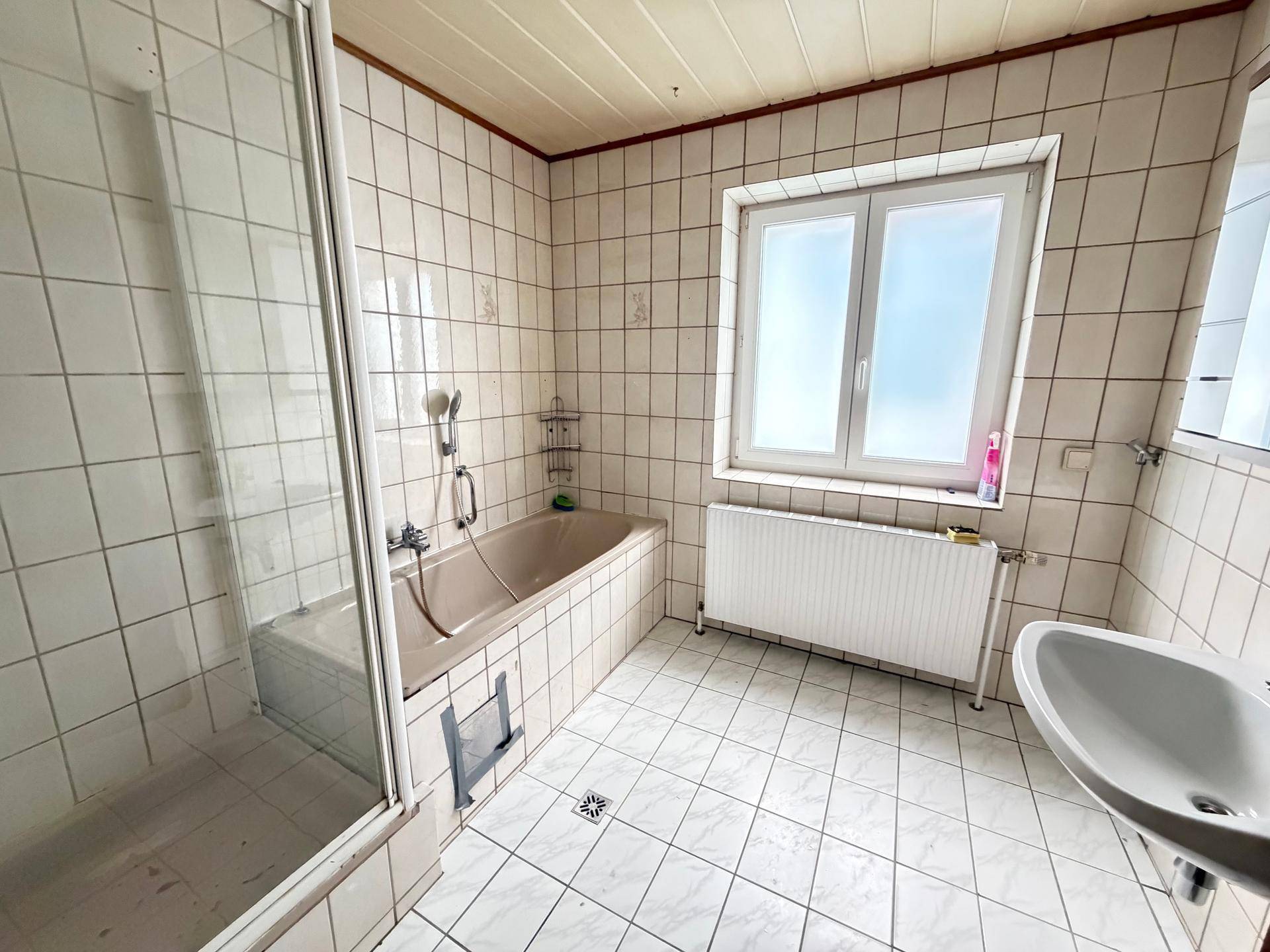 Gefliestes Badezimmer mit Badewanne, integrierter Dusche, Waschbecken und Fenster, im Stil der 80er Jahre.