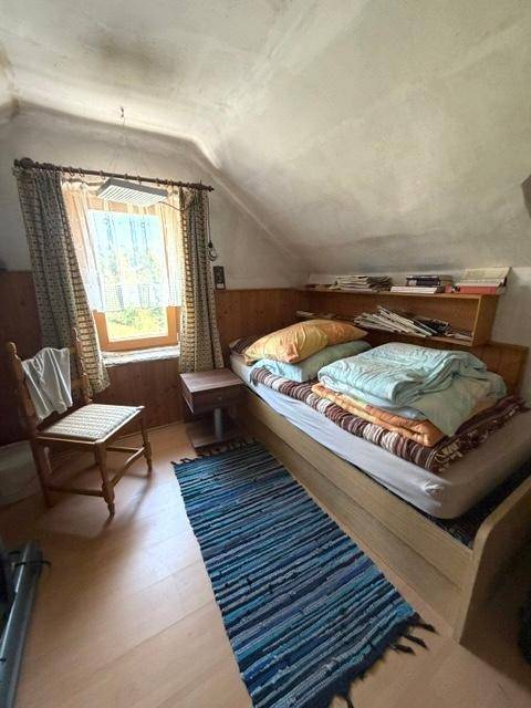 Dachschlafzimmer mit Holzbett, Teppich und schrägen Wänden, durch das Fenster fällt Licht.