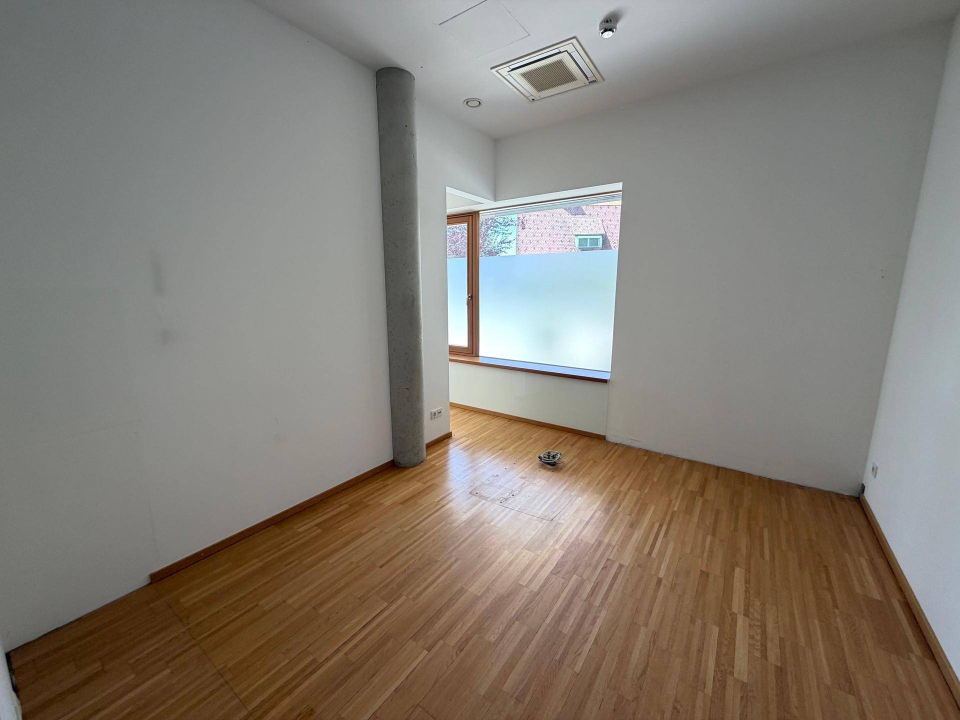 Leerer Büroraum mit Holzfußboden, weißen Wänden, einer Betonsäule und einem Fenster.
