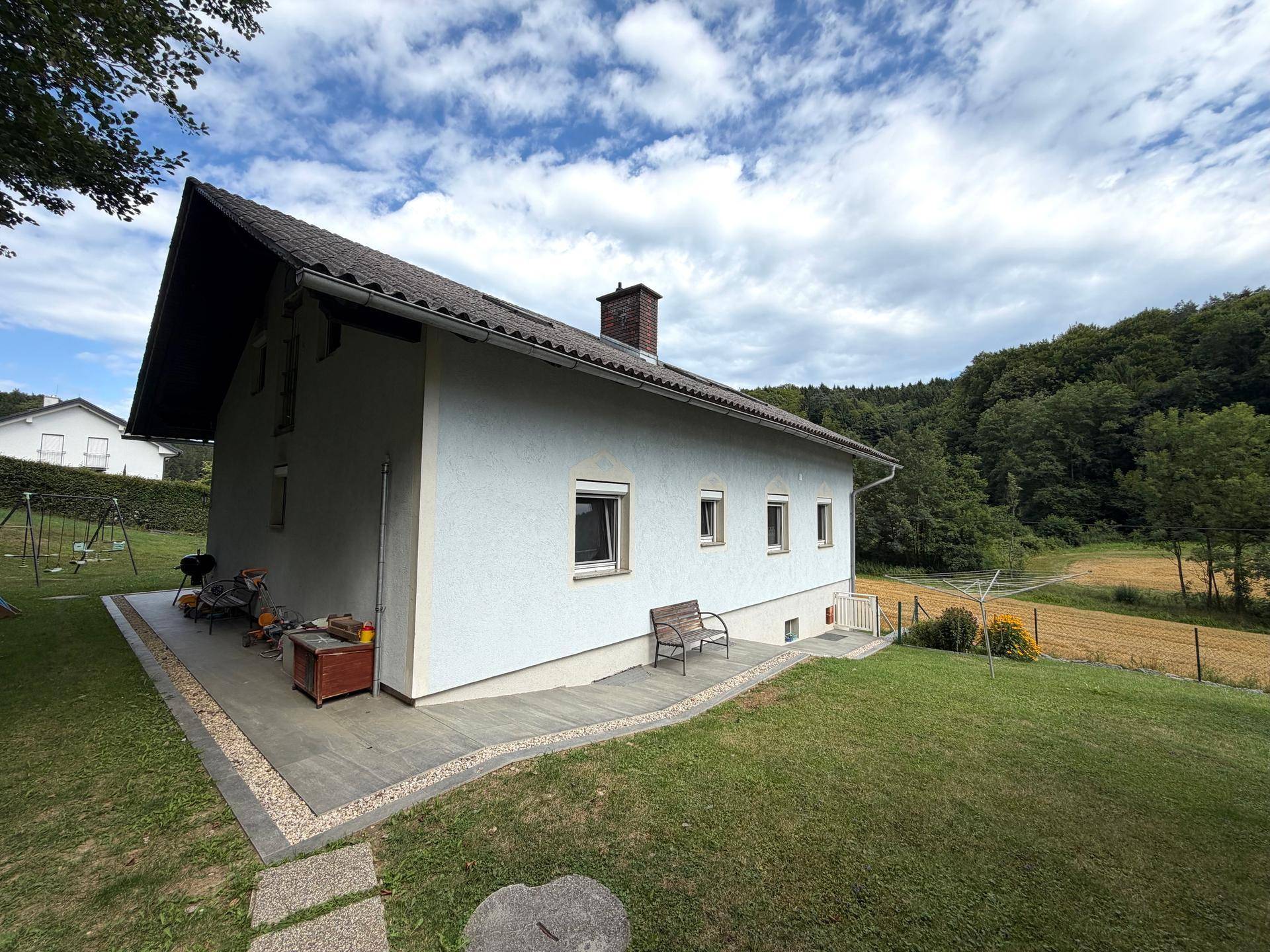Seitenansicht des Hauses mit gepflegtem Rasen und angrenzendem Feld unter blauem Himmel.