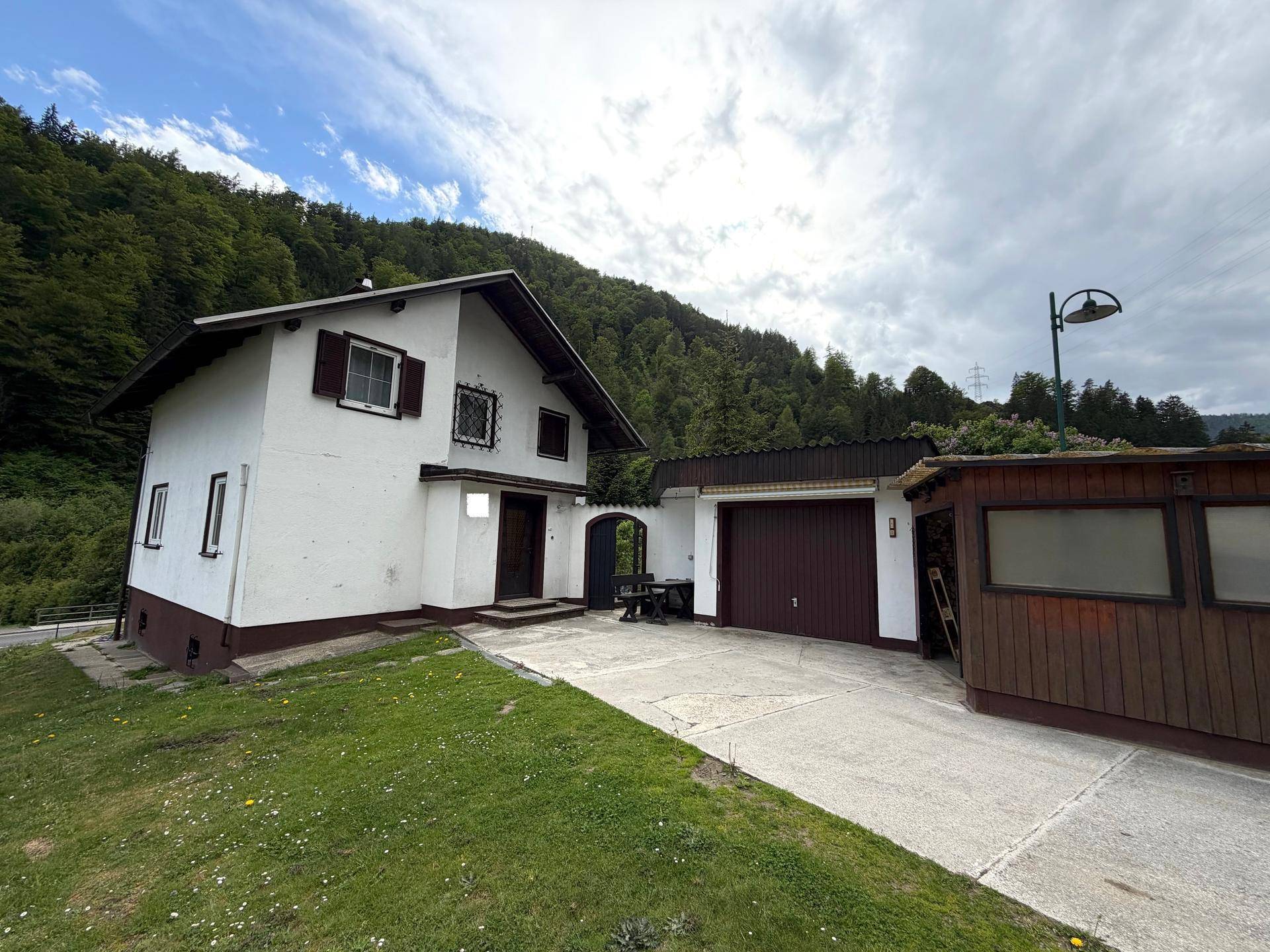Einfamilienhaus mit integrierter Garage, breiter Zufahrt und umgebender Natur in bergiger Landschaft.