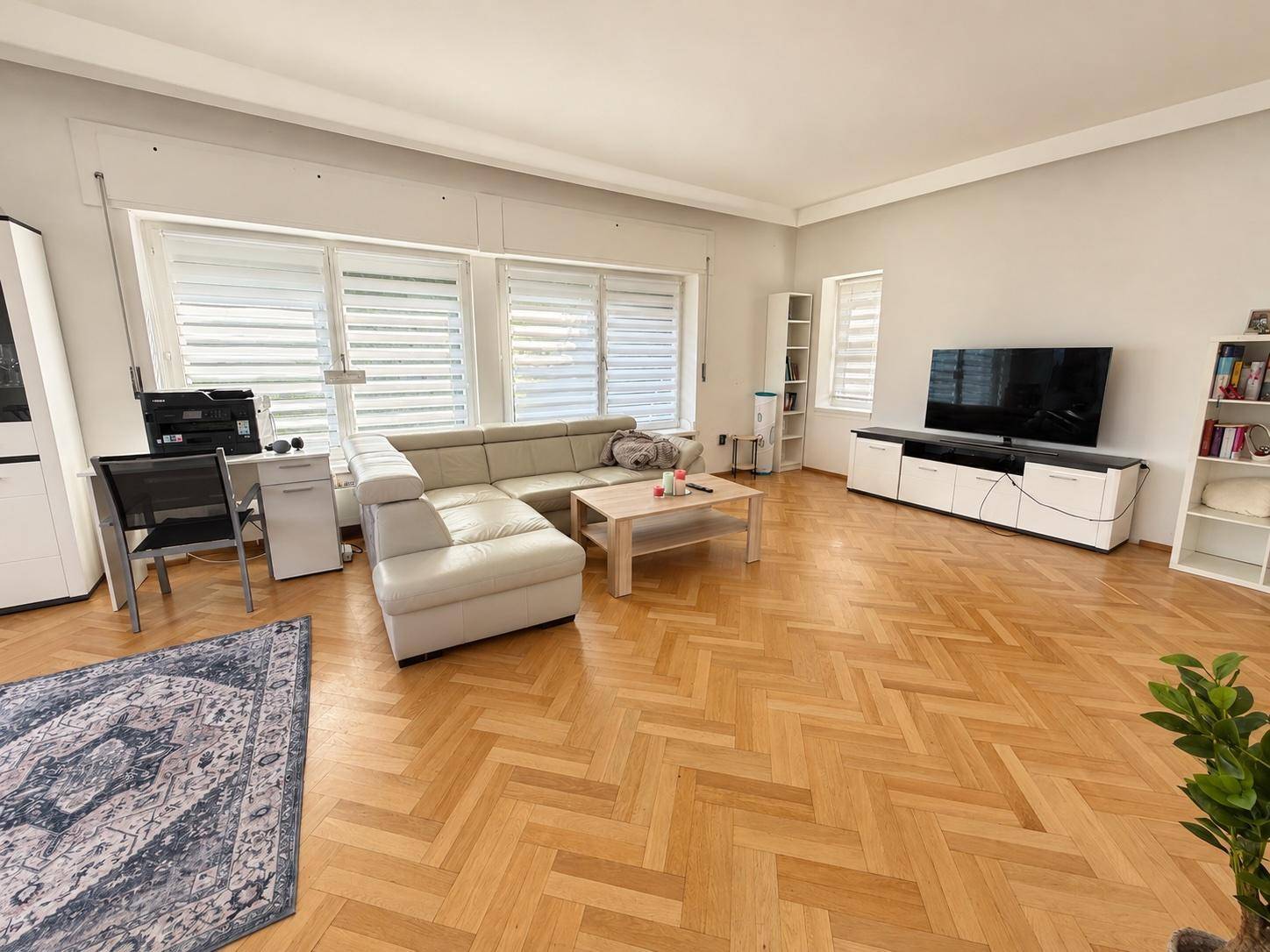 Helles Wohnzimmer mit Fischgrätparkett, großem Sofa und breiten Fenstern mit Rollläden.