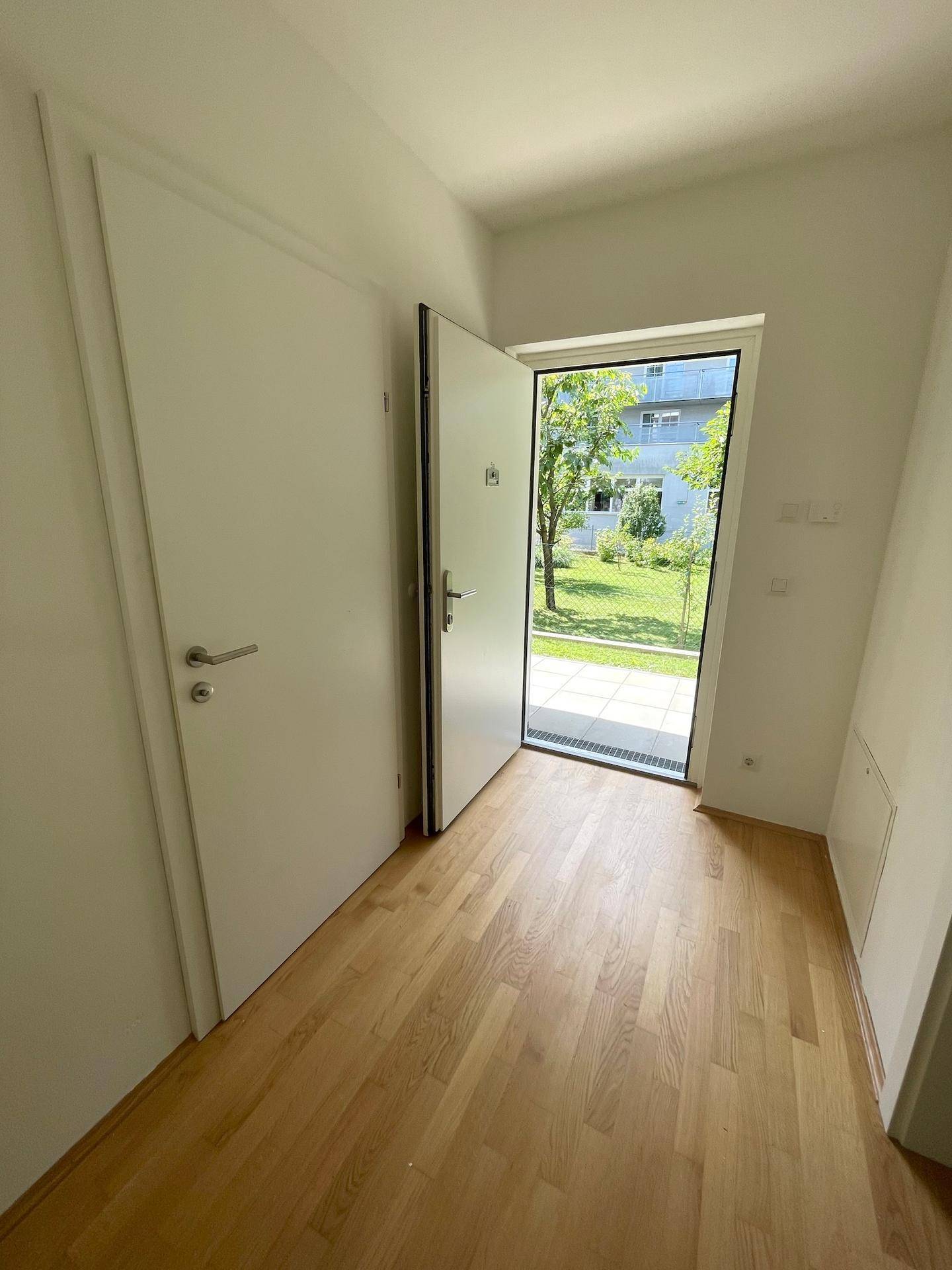 Heller Eingangsbereich mit Holzboden und offener Tür, die den Blick auf einen grünen Innenhof freigibt.