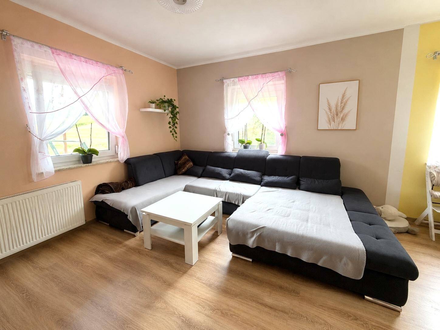 Gemütliches Wohnzimmer mit großem Ecksofa, Couchtisch und hellen Vorhängen an den Fenstern.