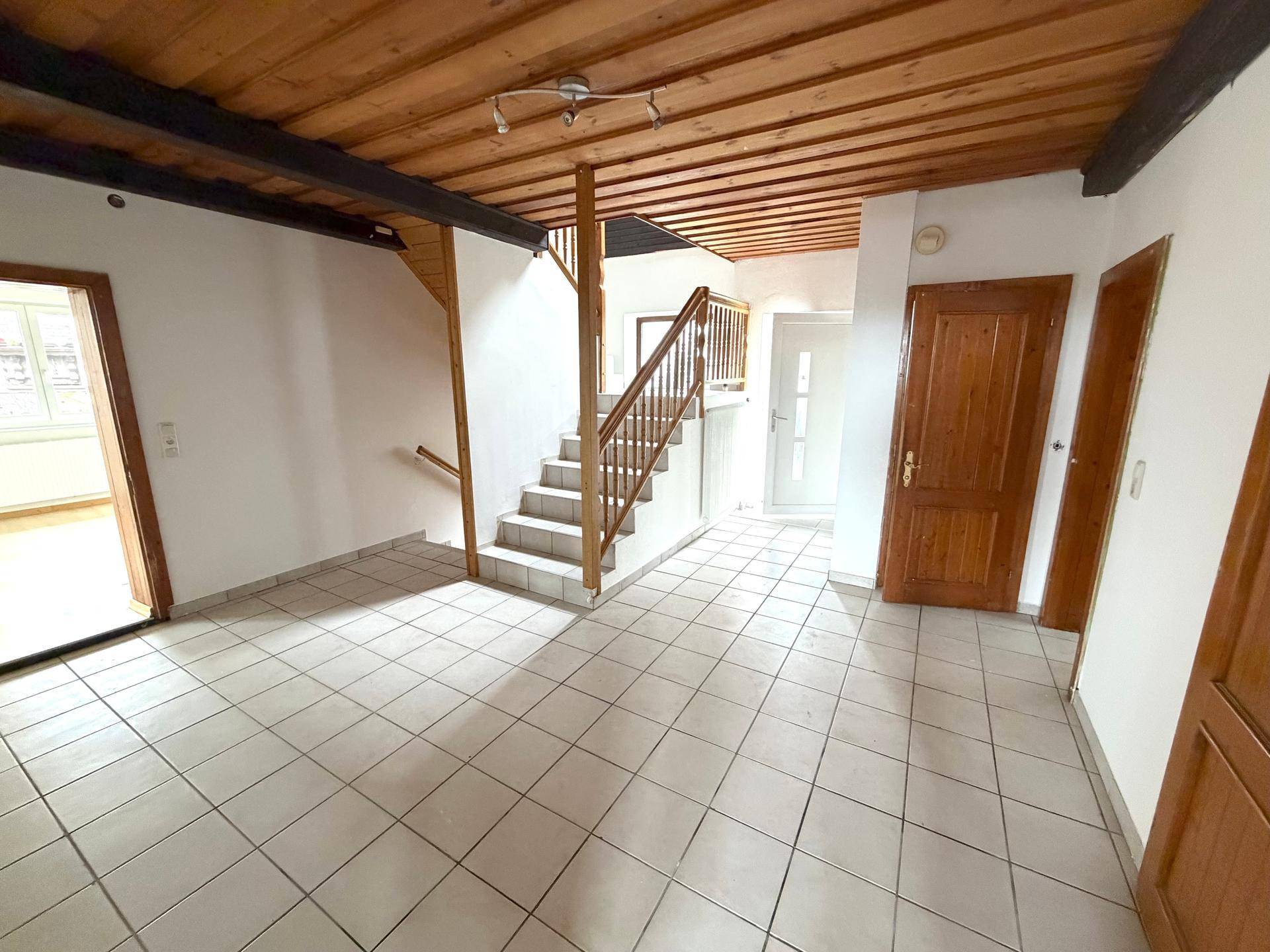 Heller Eingangsbereich mit Fliesenboden, sichtbaren Holzbalken an der Decke und offener Holztreppe.