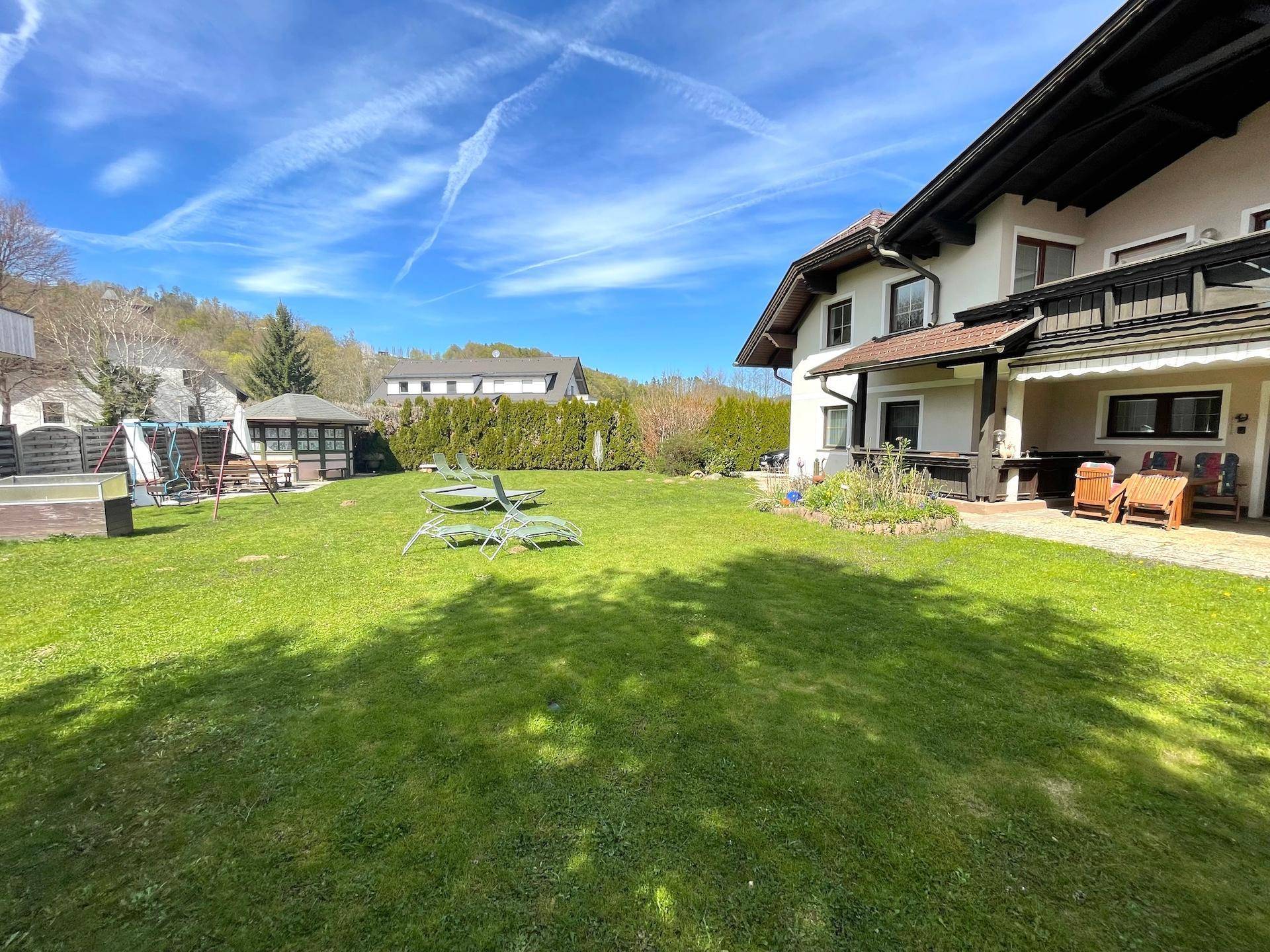 Ein weitläufiger Gartenbereich mit Rasenfläche, Spielplatz und Gartenhaus im Sonnenschein.
