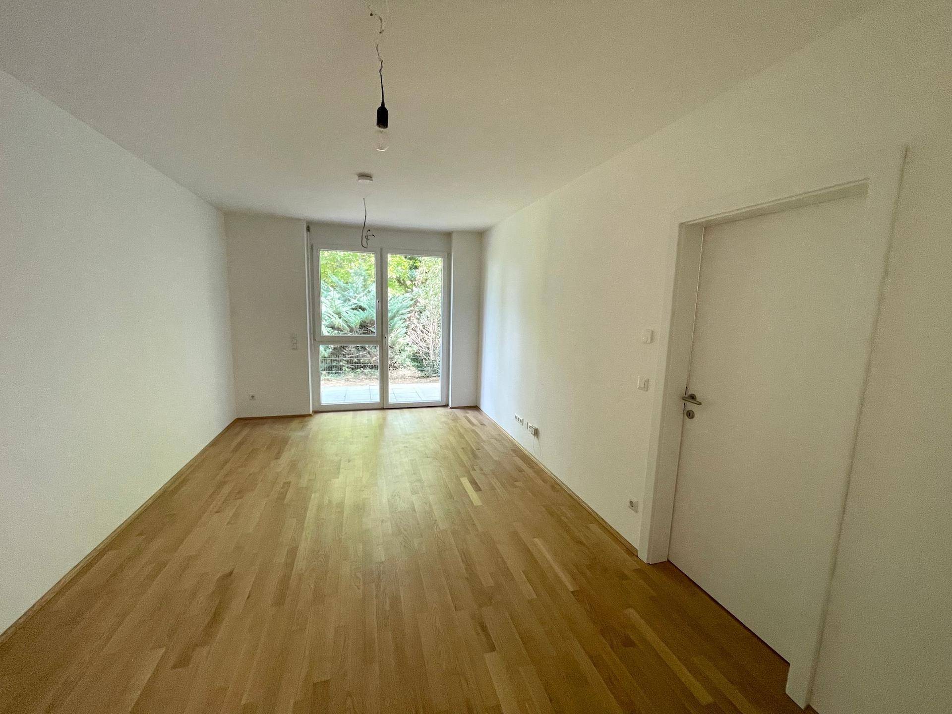 Geräumiger, heller Raum mit Holzboden und großer Glastür, die Zugang zum Außenbereich bietet.
