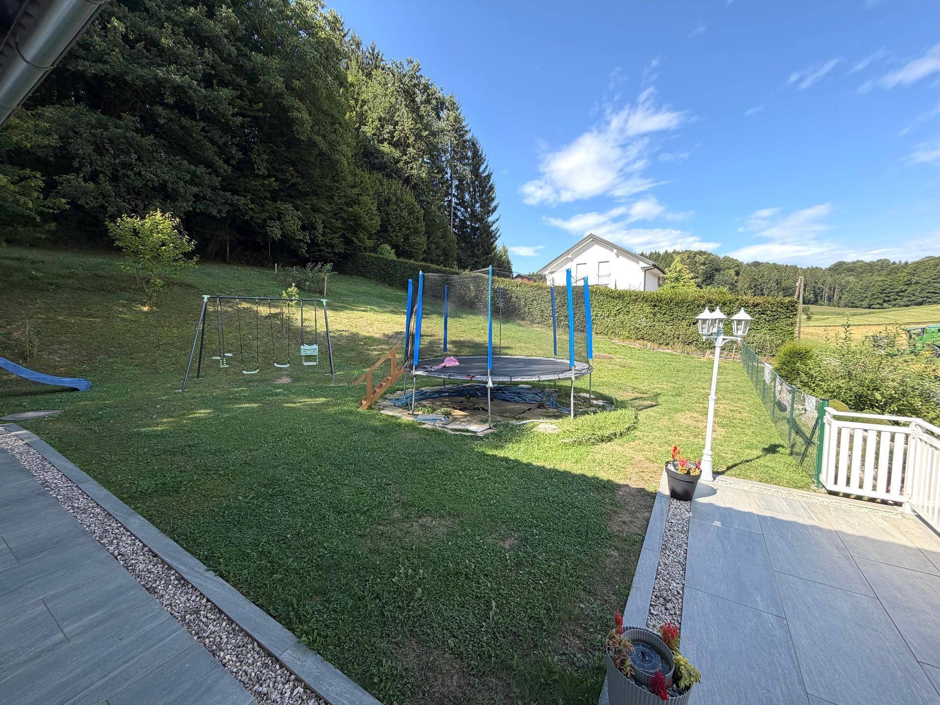 Großer Garten mit Rasenfläche, Trampolin und Schaukeln, umgeben von Bäumen und Natur.