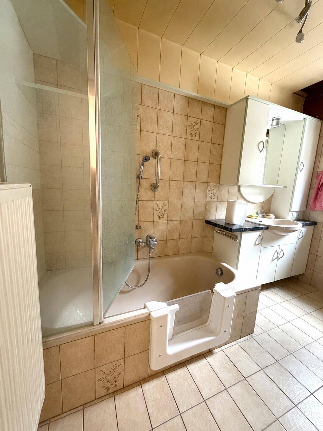 Badezimmer mit beigefarbenen Fliesen, Badewanne mit Einstiegshilfe und Waschtisch.