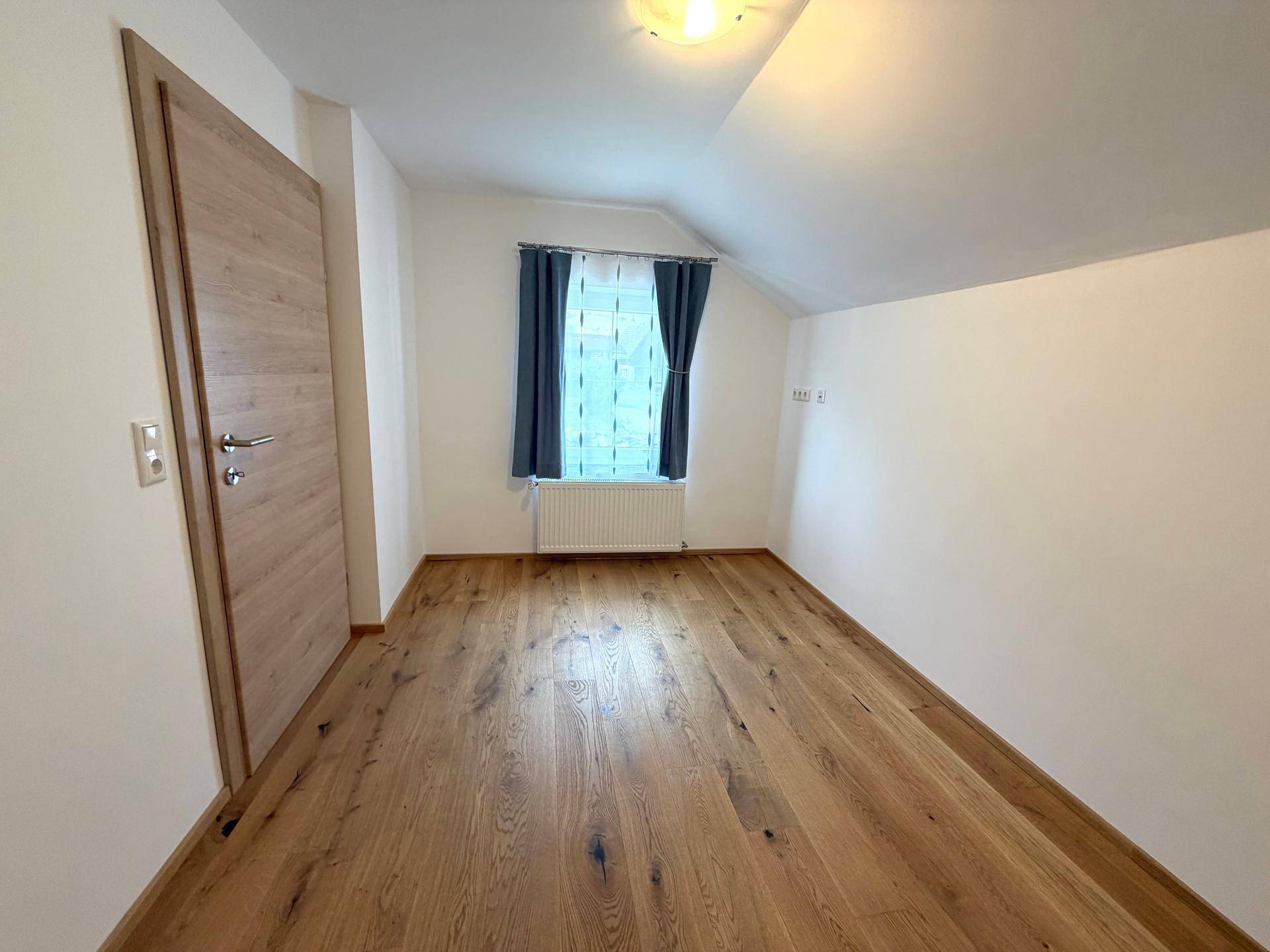 Schlafzimmer mit Holzboden, schrägen Wänden und Fenster mit Vorhängen.