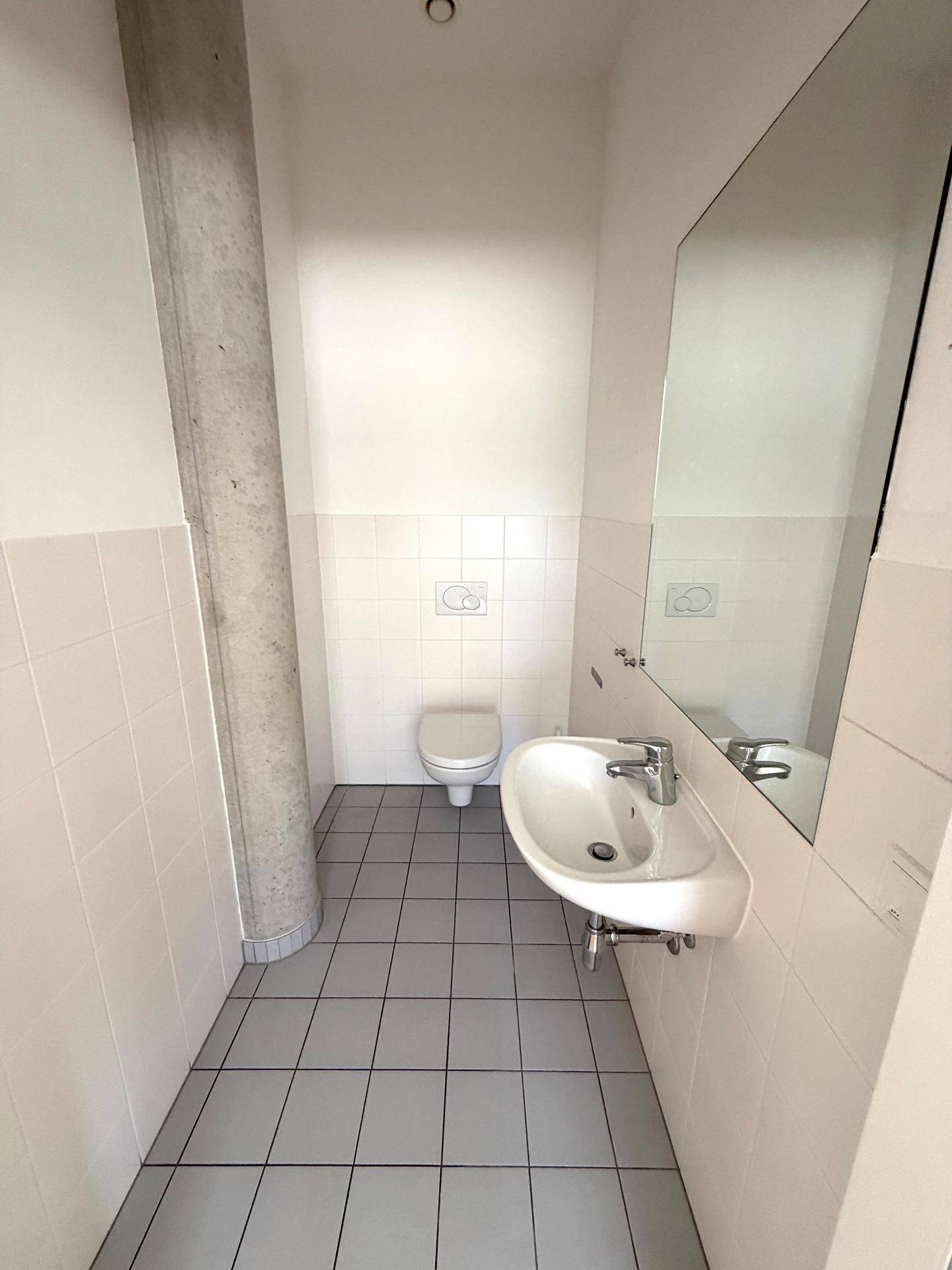 Modernes WC mit weißen Fliesen, grauem Fliesenboden, Waschbecken und großem Spiegel.