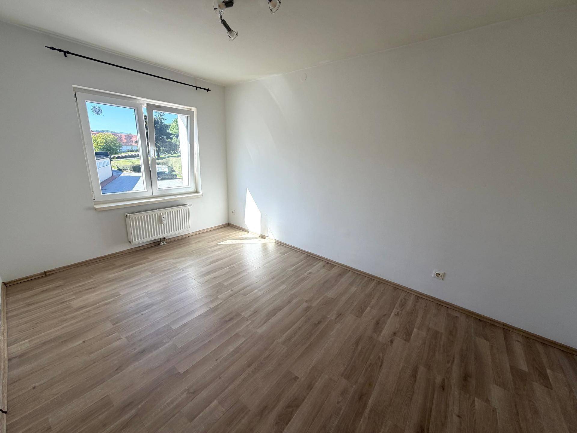 Helles Zimmer mit Laminatboden, weißen Wänden und einem Fenster mit Blick ins Grüne.