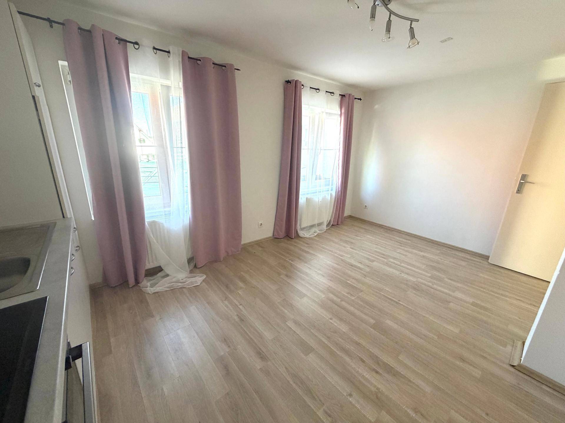 Heller Wohnbereich mit Laminatboden, weißen Wänden und zwei Fenstern mit rosa Vorhängen.