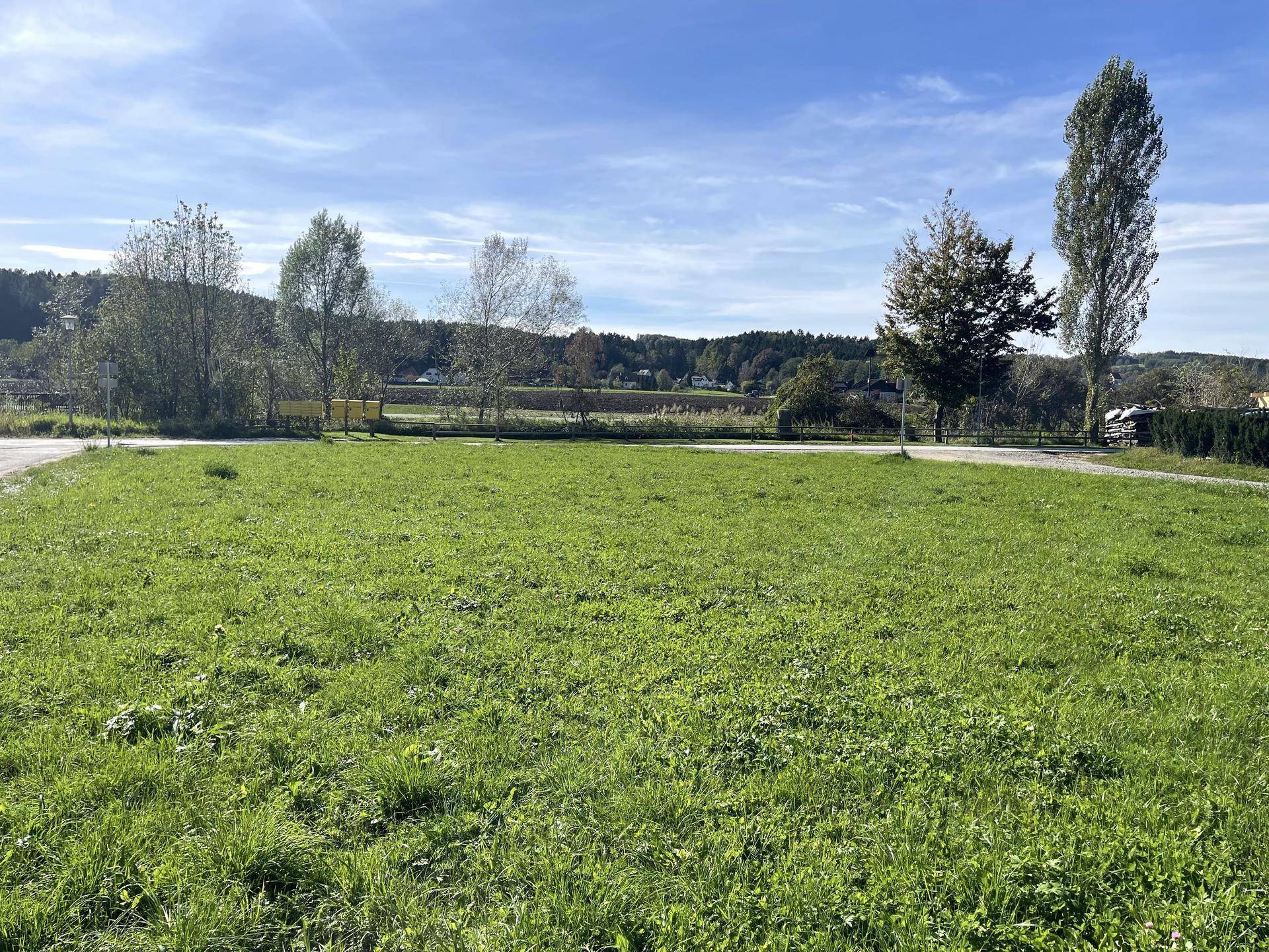 Offene Grünfläche mit Wegen und Bäumen, umgeben von ländlicher Landschaft unter blauem Himmel.