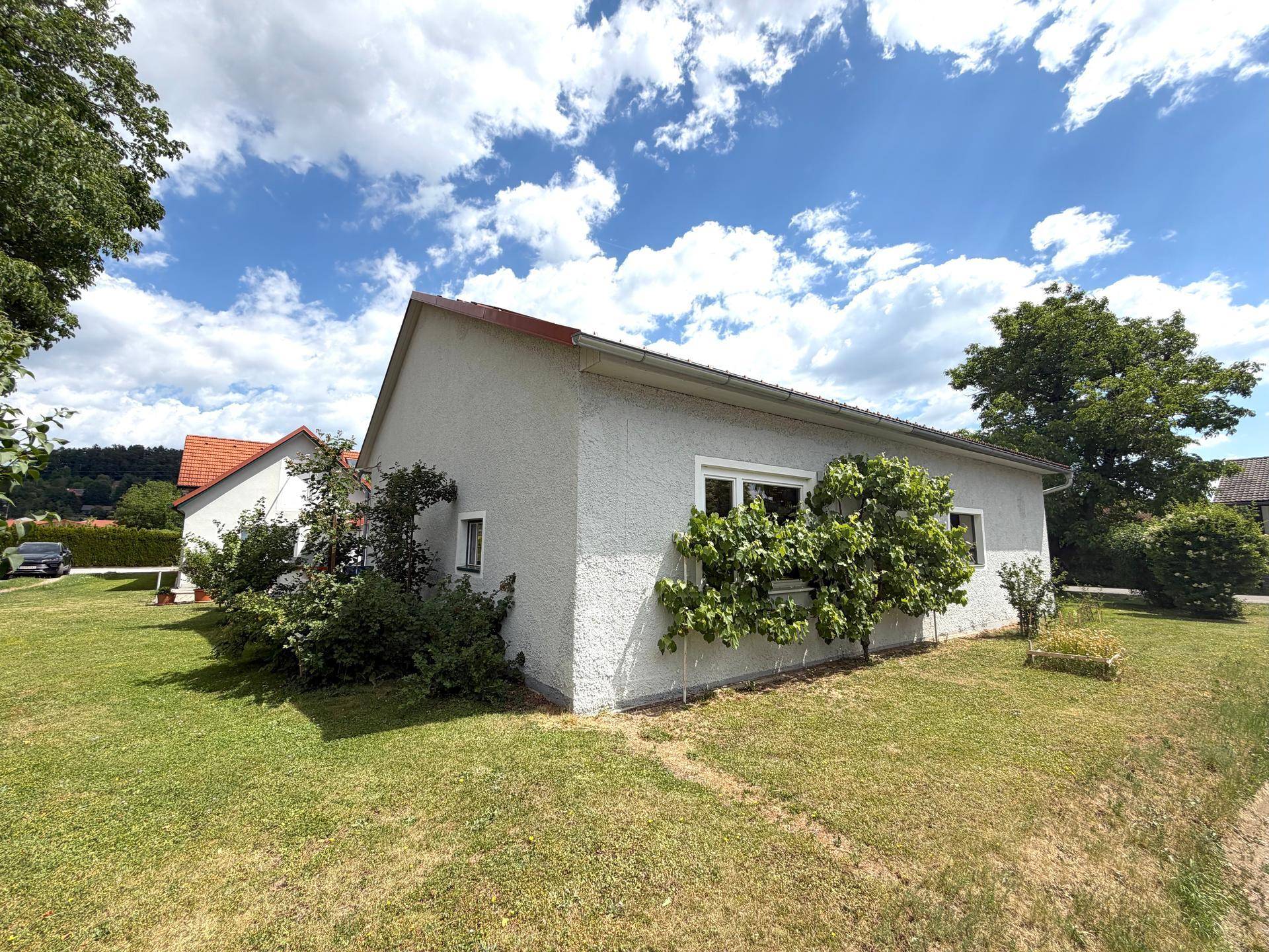 Seitenansicht des Hauses mit heller Fassade, rotem Ziegeldach und umgebendem Grünbereich.