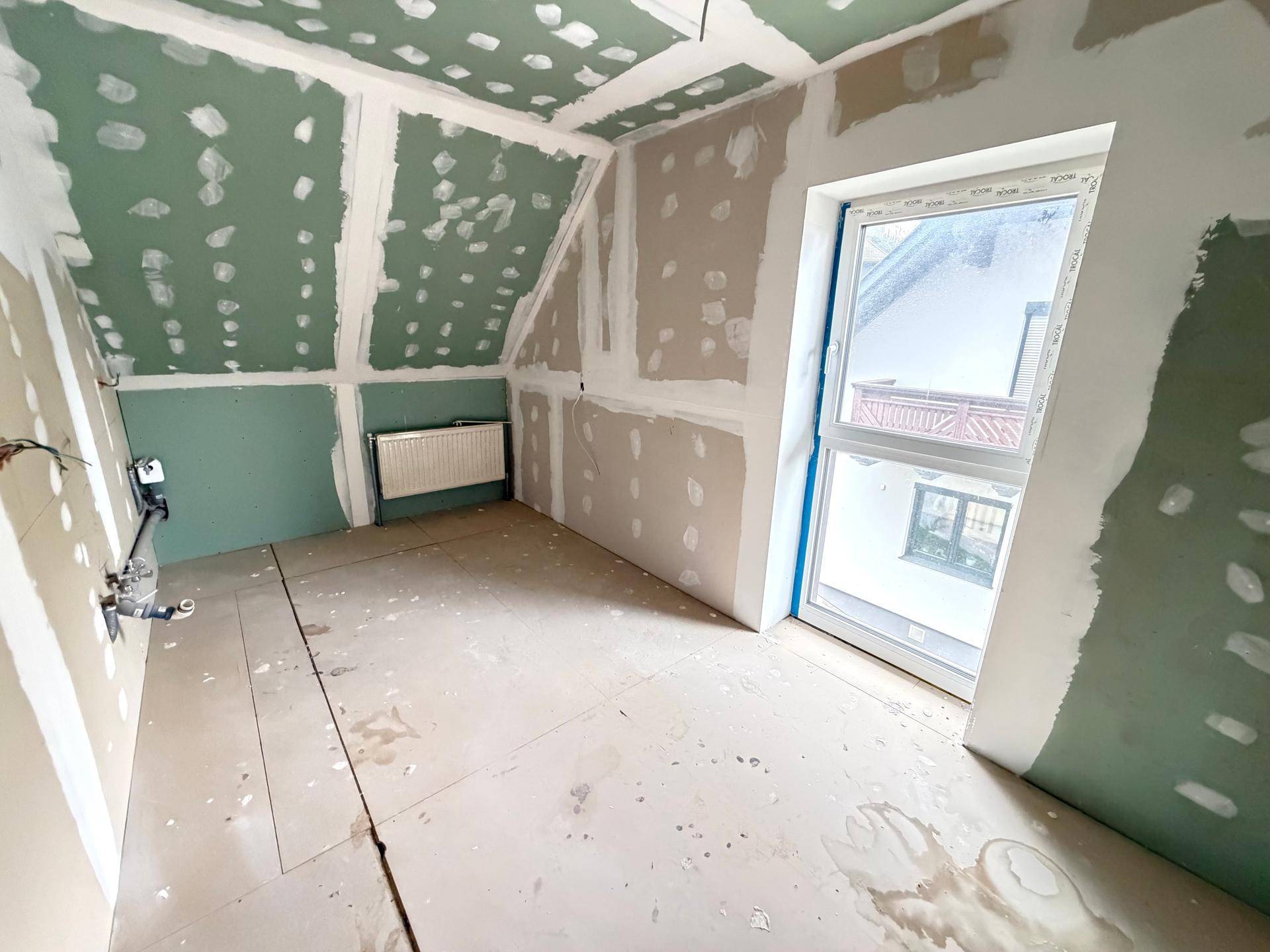 Dachgeschosszimmer im Rohbau mit schräger Decke, Gipskartonwänden und Fenster, bereit für den Innenausbau.