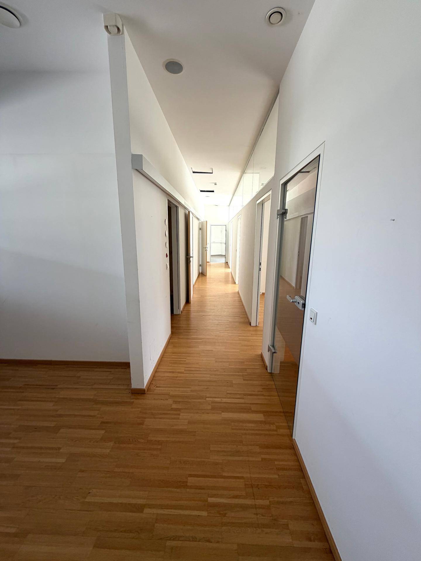 Langer, heller Flur mit Holzfußboden und weißen Wänden, der zu mehreren Büroräumen führt.