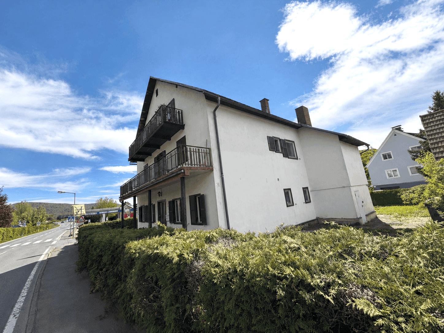 Weißes Wohnhaus mit mehreren Balkonen und dunklen Fensterläden an einer Straße gelegen.