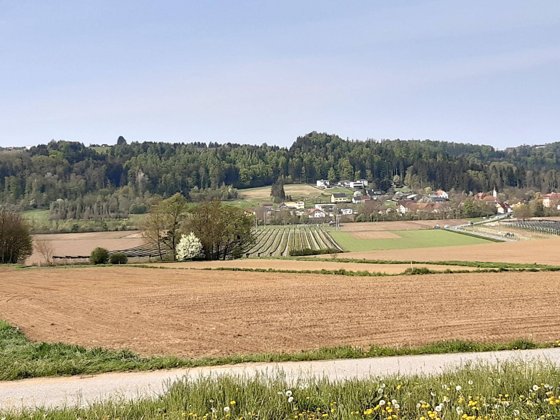 Ländliche Landschaft mit braunen Feldern, grünen Hügeln und einem kleinen Dorf am Horizont.