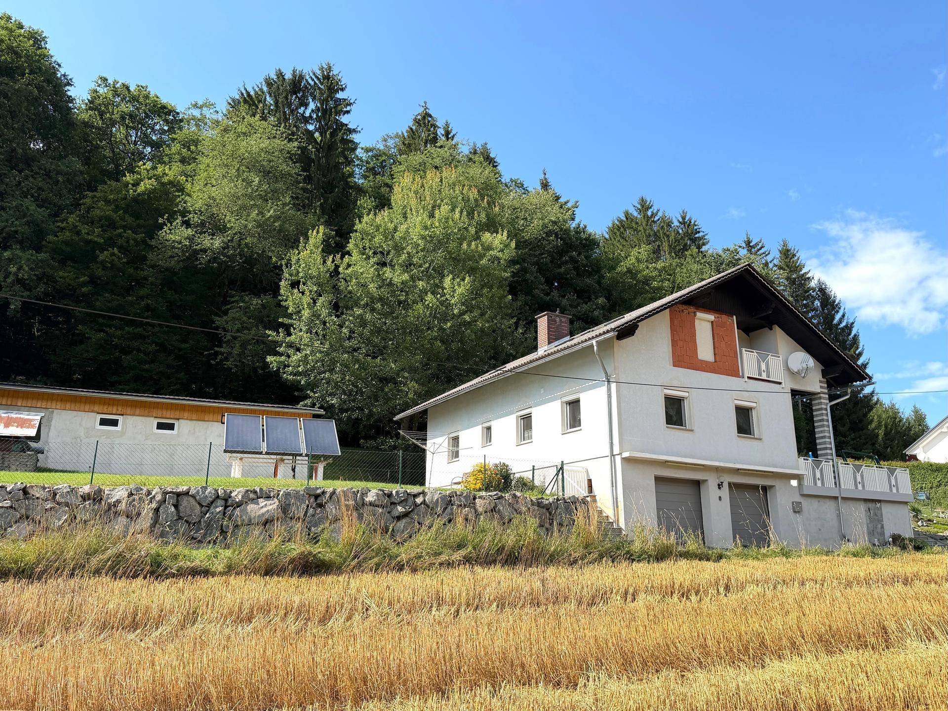 Ansicht des Hauses und eines Nebengebäudes mit Solaranlage, umgeben von Feldern und Wald.