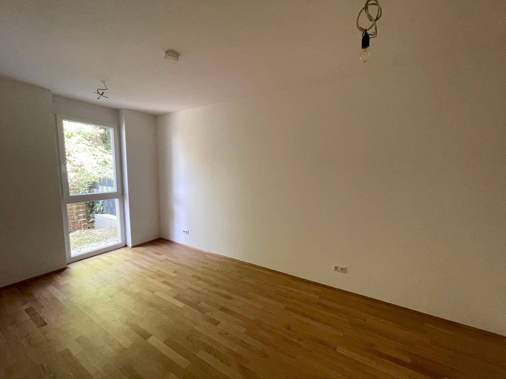 Lichtdurchfluteter Raum mit Holzboden und hoher Fenstertür, die viel Tageslicht hereinlässt.