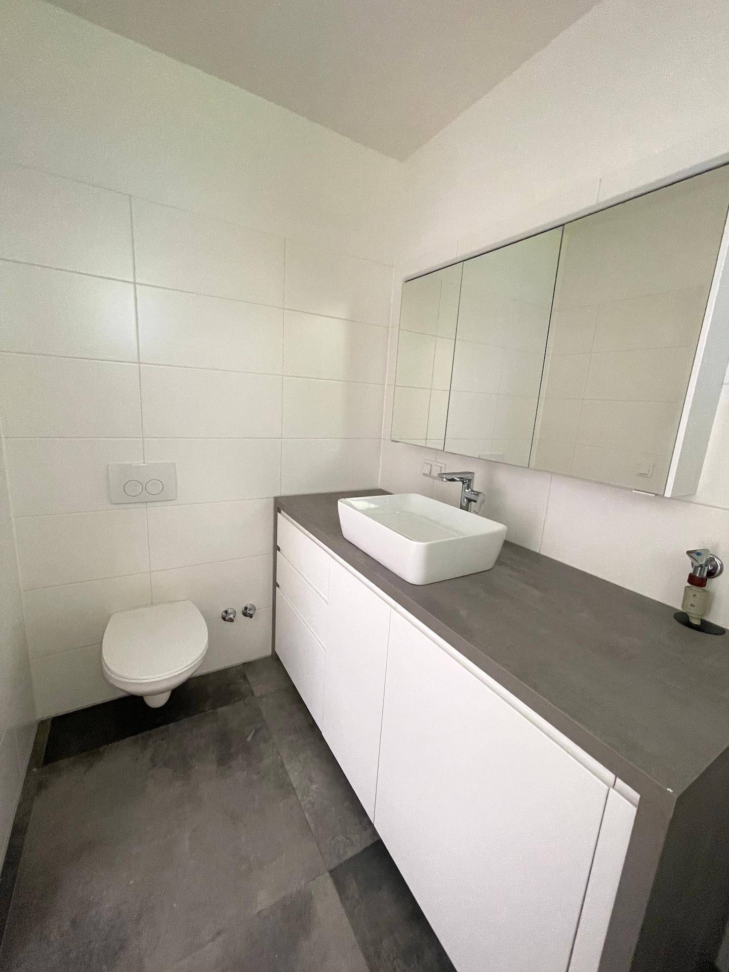 Modernes Badezimmer mit weißen Fliesen, dunklem Boden, elegantem Waschtisch und wandhängendem WC.