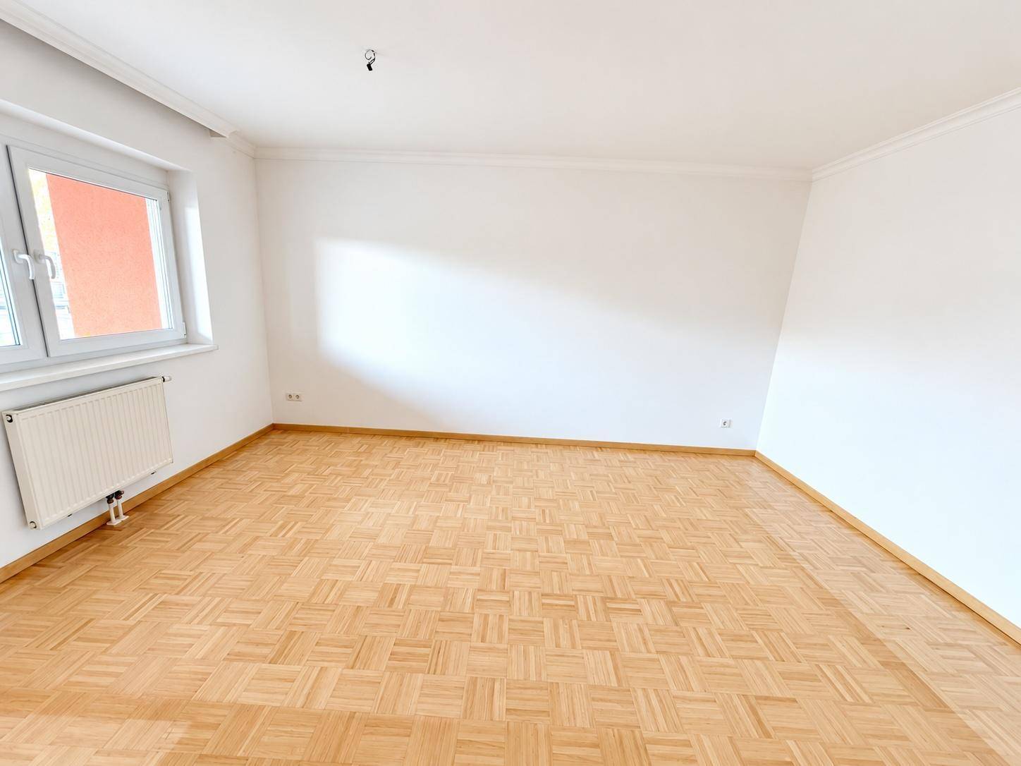 Geräumiger Wohnbereich mit hellem Parkett und großen Fenstern, die viel Tageslicht hereinlassen.