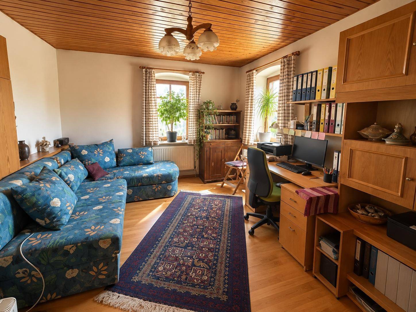 Geräumiges Wohnzimmer mit blauer Eckcouch, Holzdecke und einem Arbeitsplatz am Fenster.