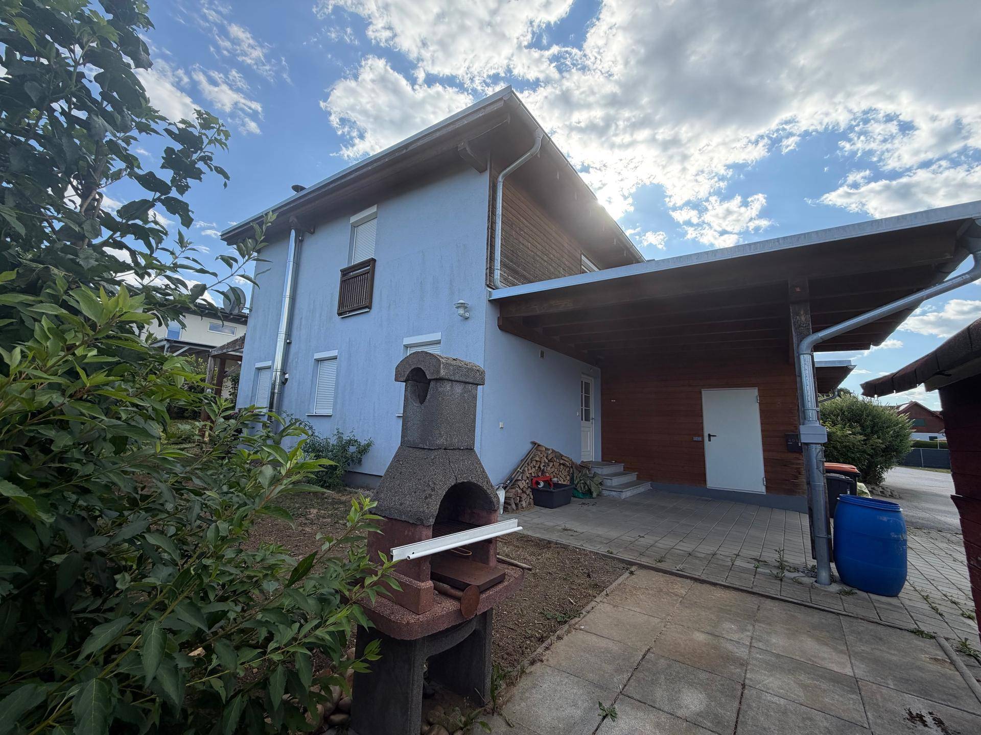 Hausansicht mit Carport, Außenkamin und gepflasterter Zufahrt unter blauem Himmel.
