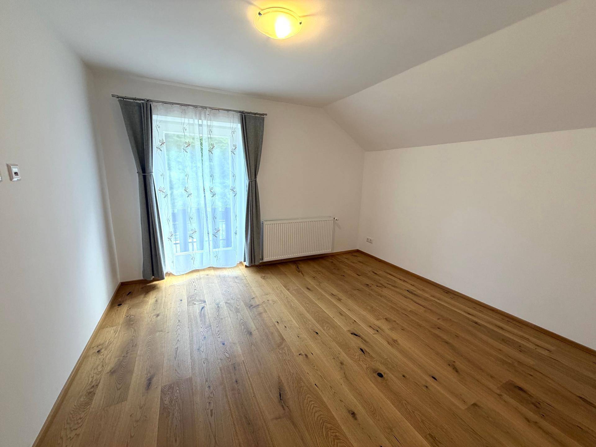 Helles Schlafzimmer mit Holzboden, schrägen Wänden und Fenster mit Vorhängen.