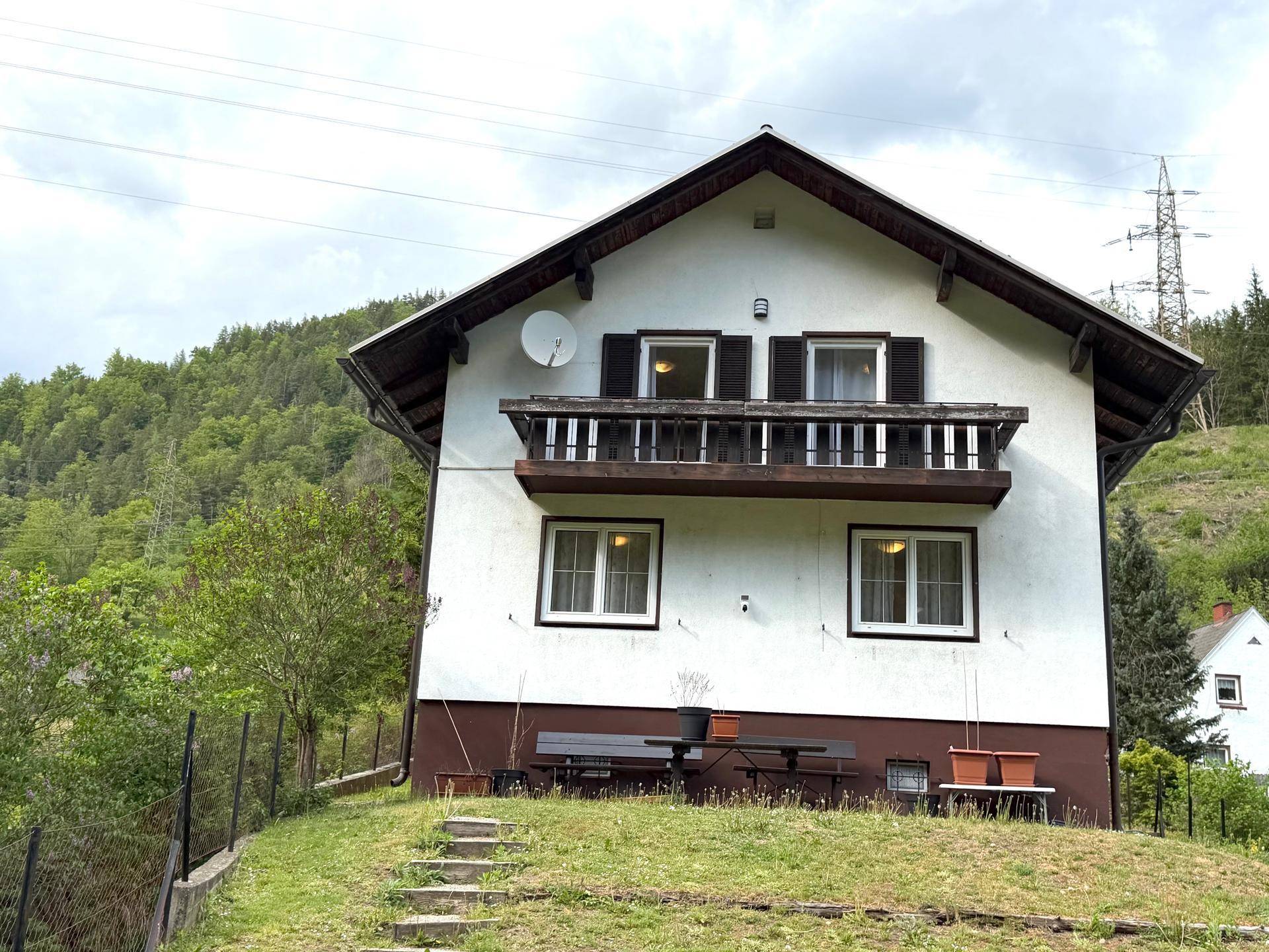 Weißes Einfamilienhaus mit Satteldach, Balkon und Blick auf bewaldete Hügel in ländlicher Umgebung.