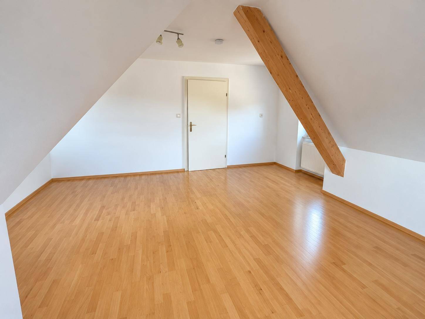 Geräumiges Zimmer im Dachgeschoss mit sichtbarem Holzbalken, Schrägen und hellem Parkettboden.