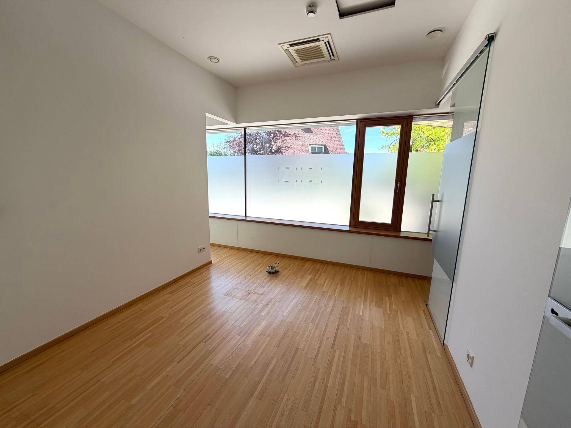Heller Büroraum mit Holzfußboden, großen Fenstern und einer modernen Glasschiebetür.