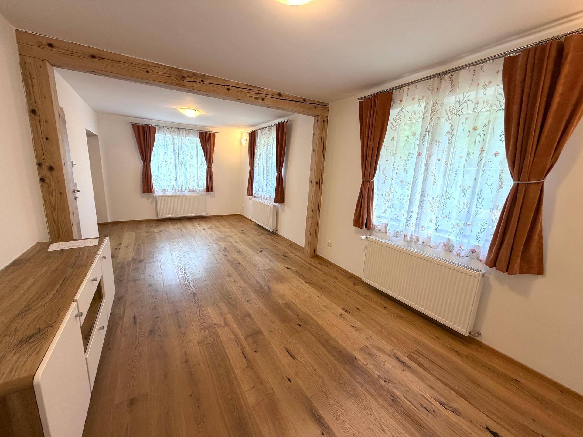 Großes Wohnzimmer mit Holzbalken, Parkettboden und mehreren Fenstern mit Vorhängen.