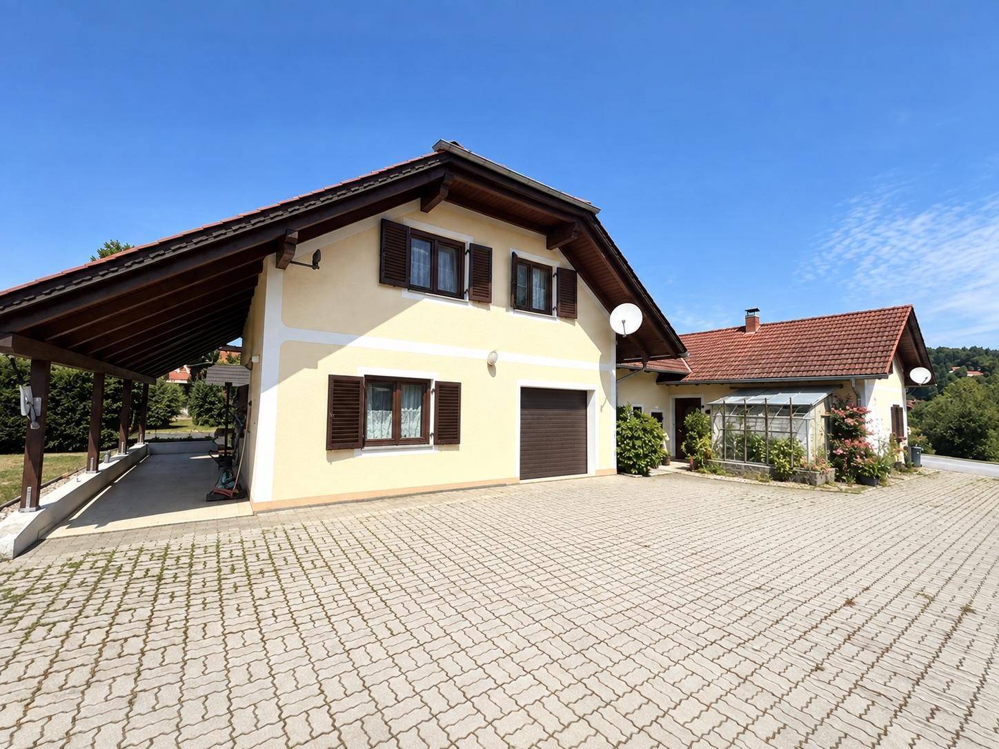 Traditionelles Haus mit gelber Fassade, Carport und gepflasterter Auffahrt unter blauem Himmel.
