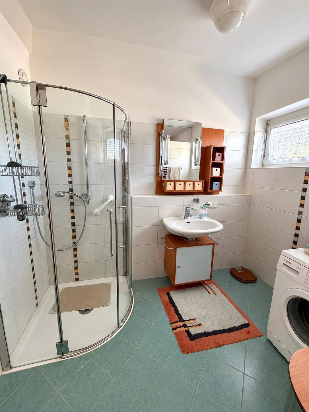 Badezimmer mit Eckdusche, Waschbecken, Spiegelschrank und Waschmaschine, hell gefliest.