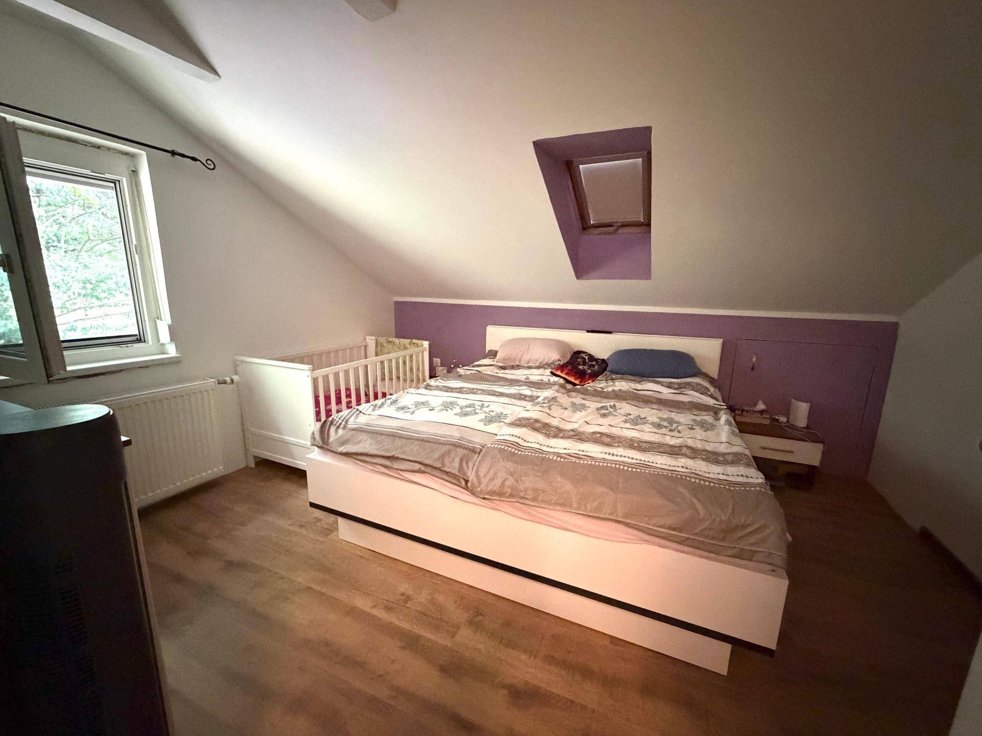 Schlafzimmer unter der Dachschräge mit Doppelbett und Dachfenster, in Lila-Tönen.