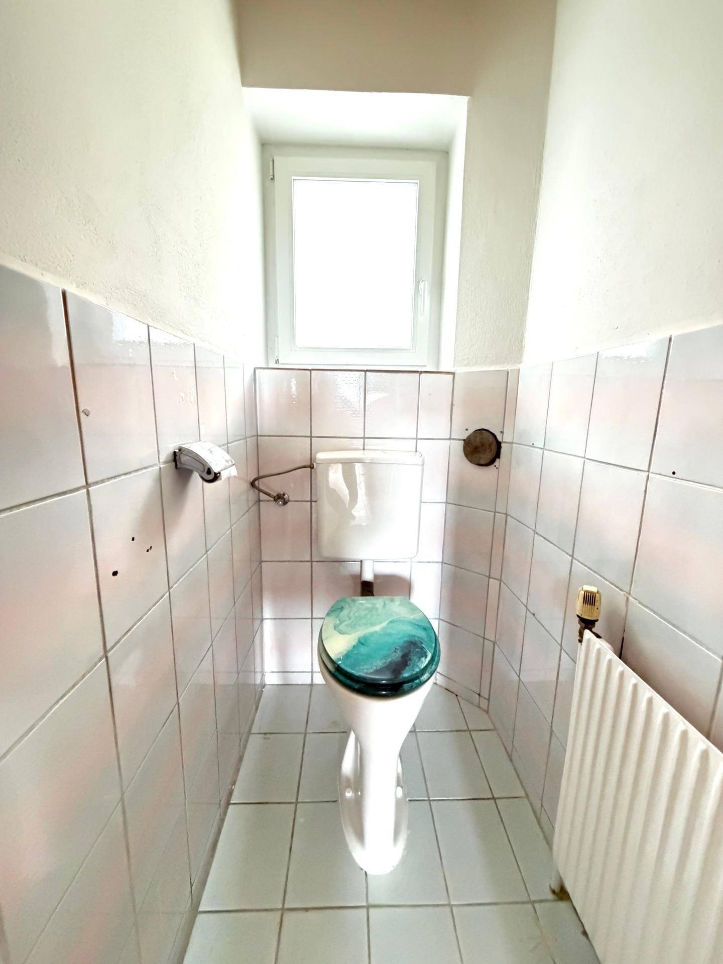 Separate Toilette mit Fenster, hellen Fliesen und einem farbigen Toilettensitz, funktional und sauber.