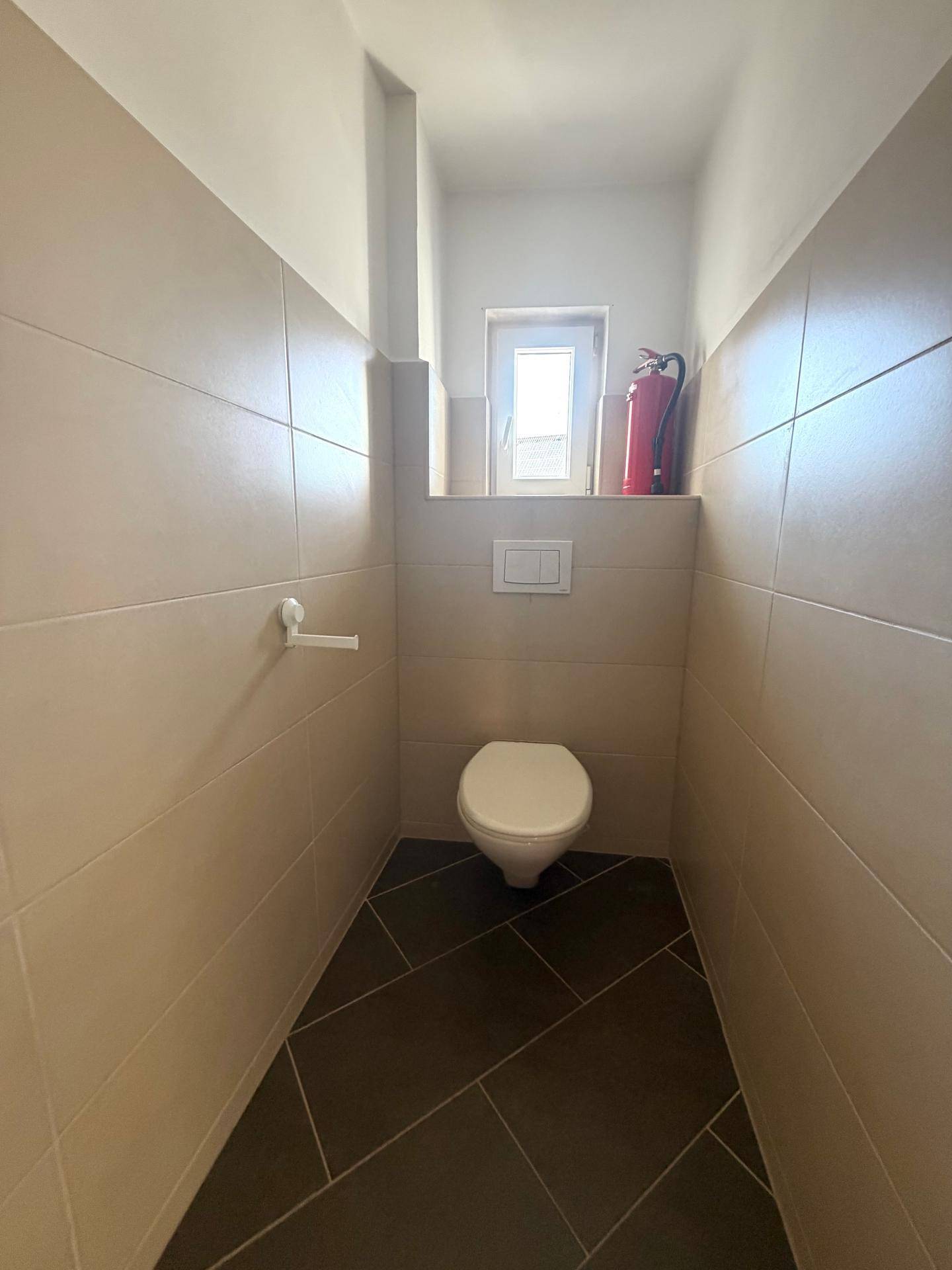 Separates WC mit Wand-WC, kleinem Fenster und beigefarbenen Wandfliesen.